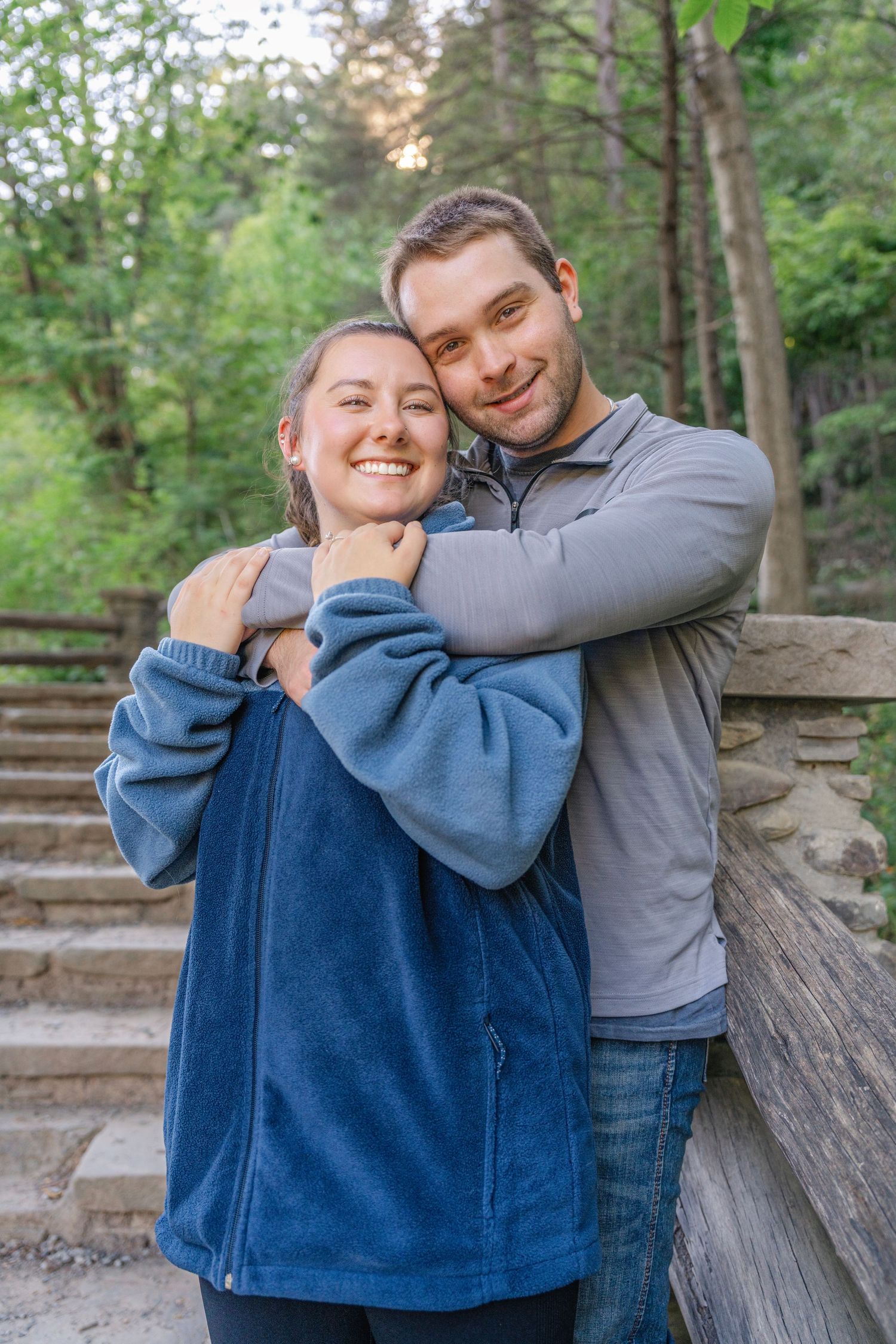 A couple shares a tender moment outdoors in a wooded setting, wearing blue and gray casual attire.