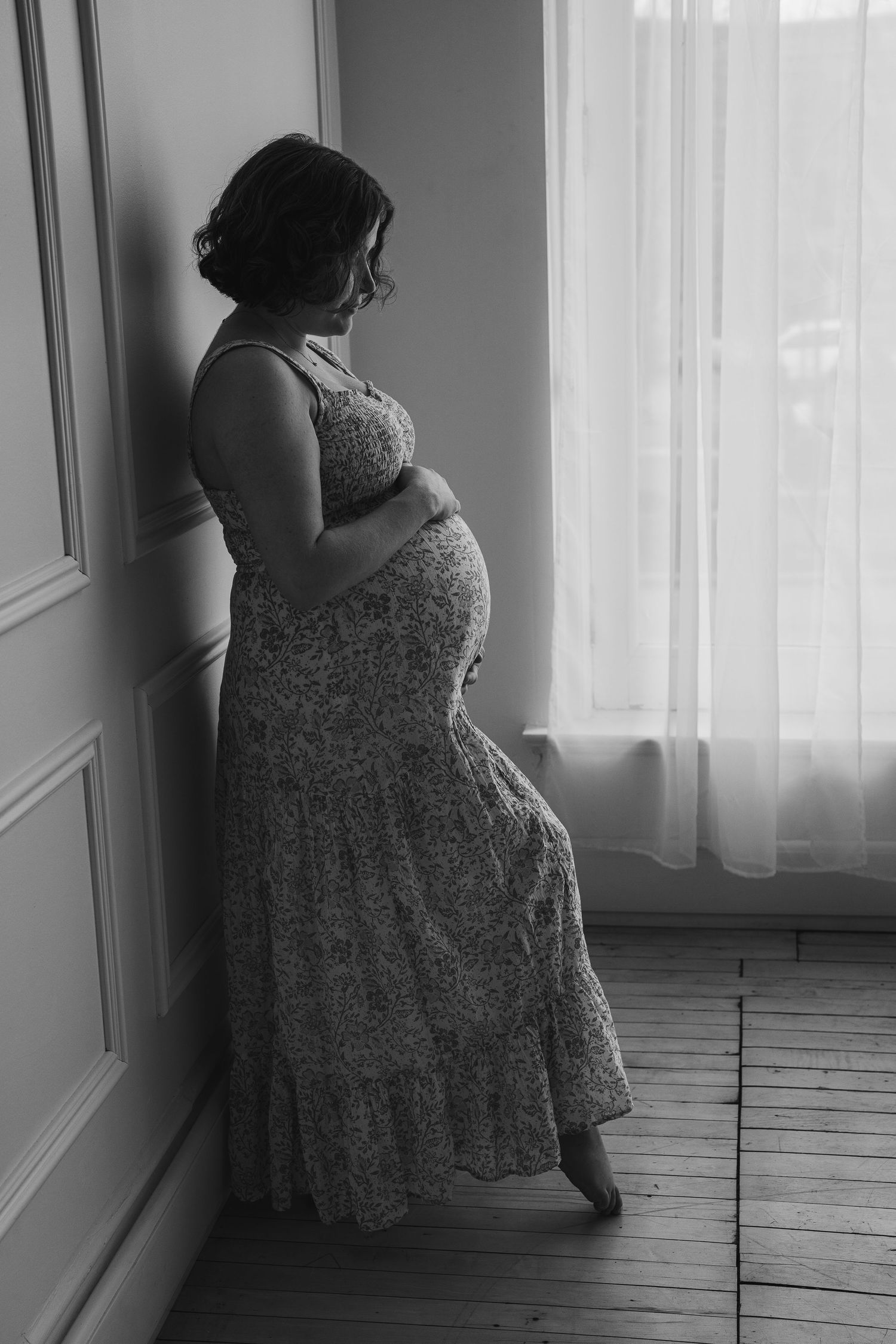 A silhouetted pregnant woman in a lace dress stands by a window in black and white, creating a dreamy maternity portrait.