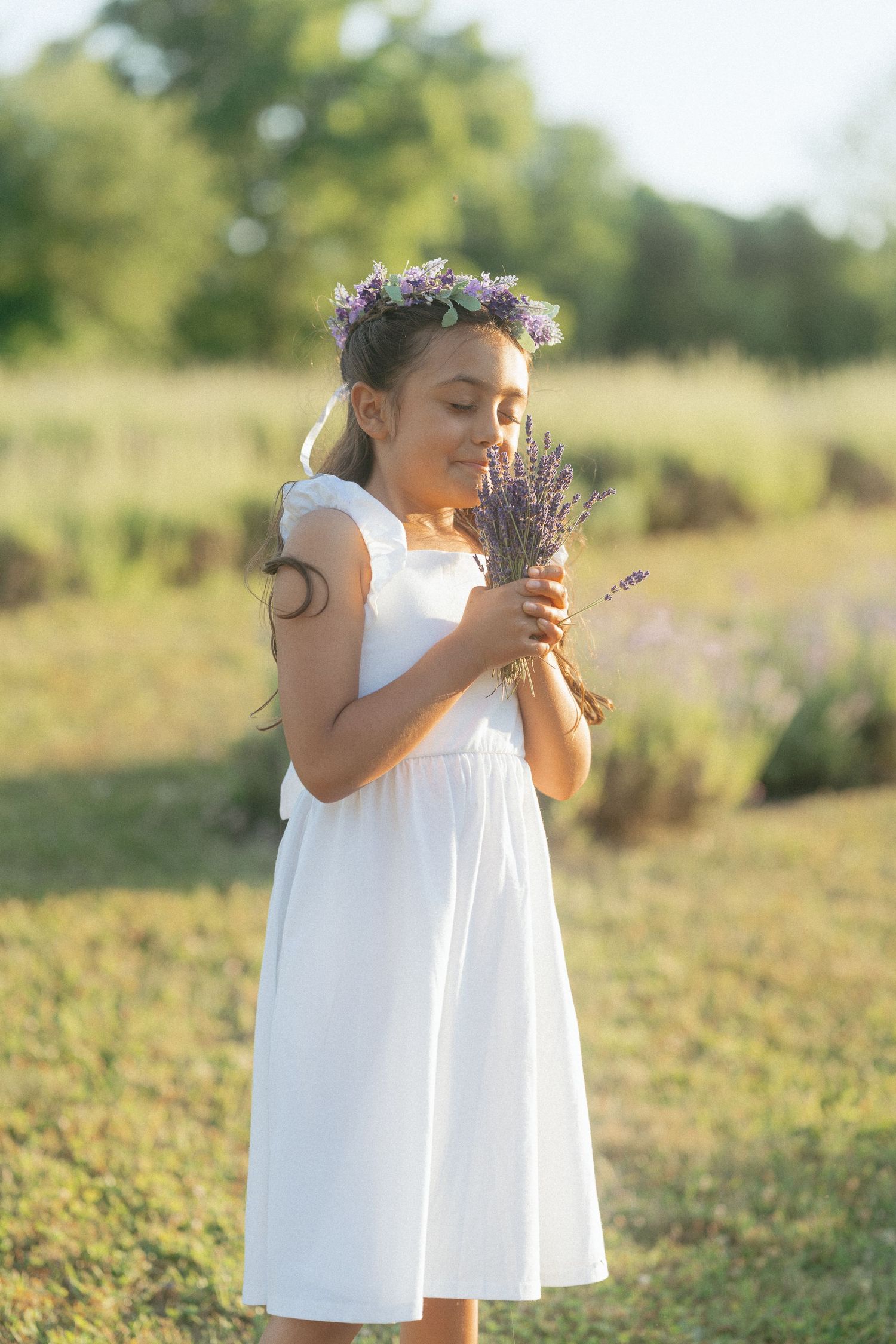 A figure in a white summer dress stands in a sunlit meadow holding a bouquet of wildflowers against a soft blurred background.