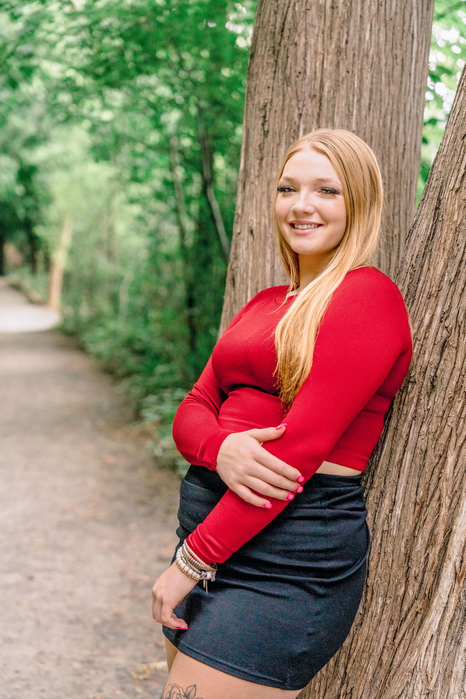 Person in red and black outfit leans against large tree trunk on forest path with natural lighting.