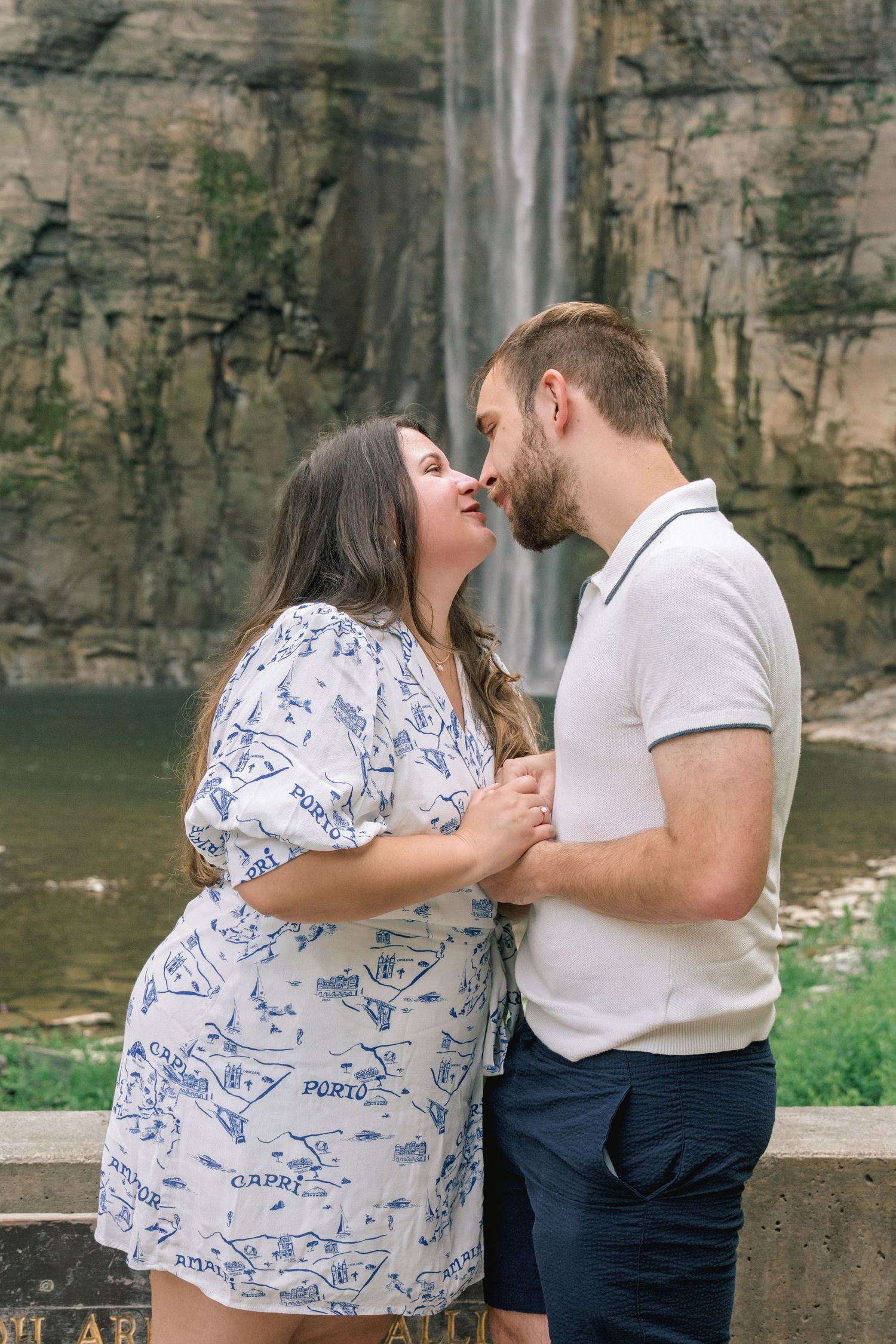 A couple shares a tender moment in front of a scenic waterfall and rocky cliffs.