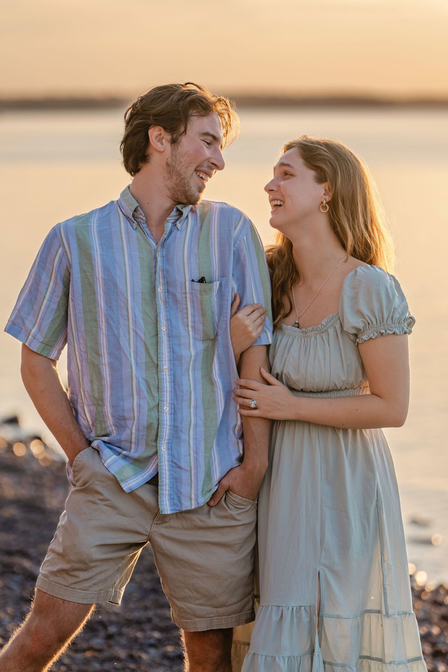 A romantic couple embraces on a beach at sunset wearing light colored clothing.