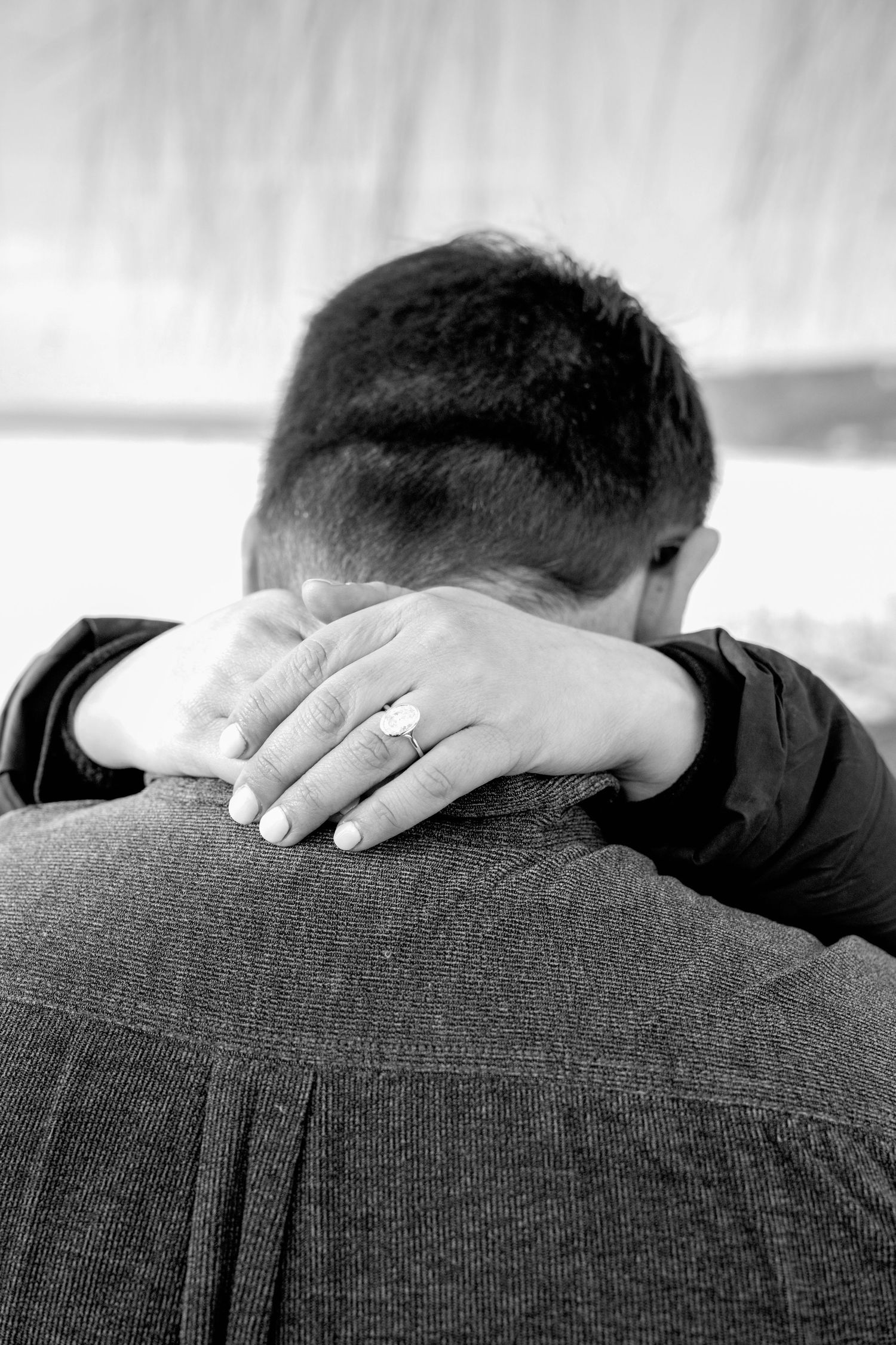 Black and white close-up of woman's hand with engagement ring wrapped around man's neck in a loving embrace.