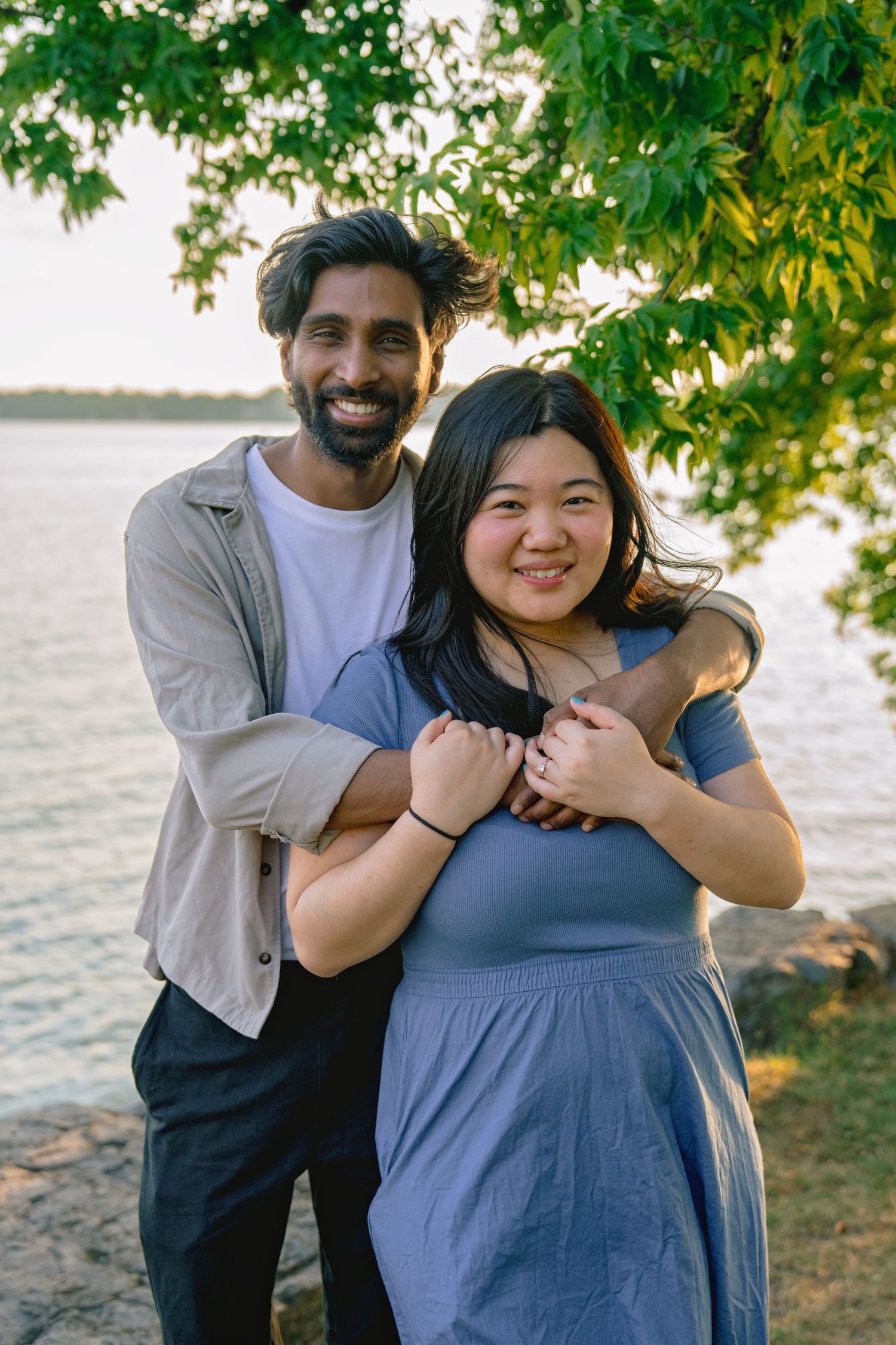A smiling couple embraces outdoors near a lake, surrounded by green trees in warm evening light.