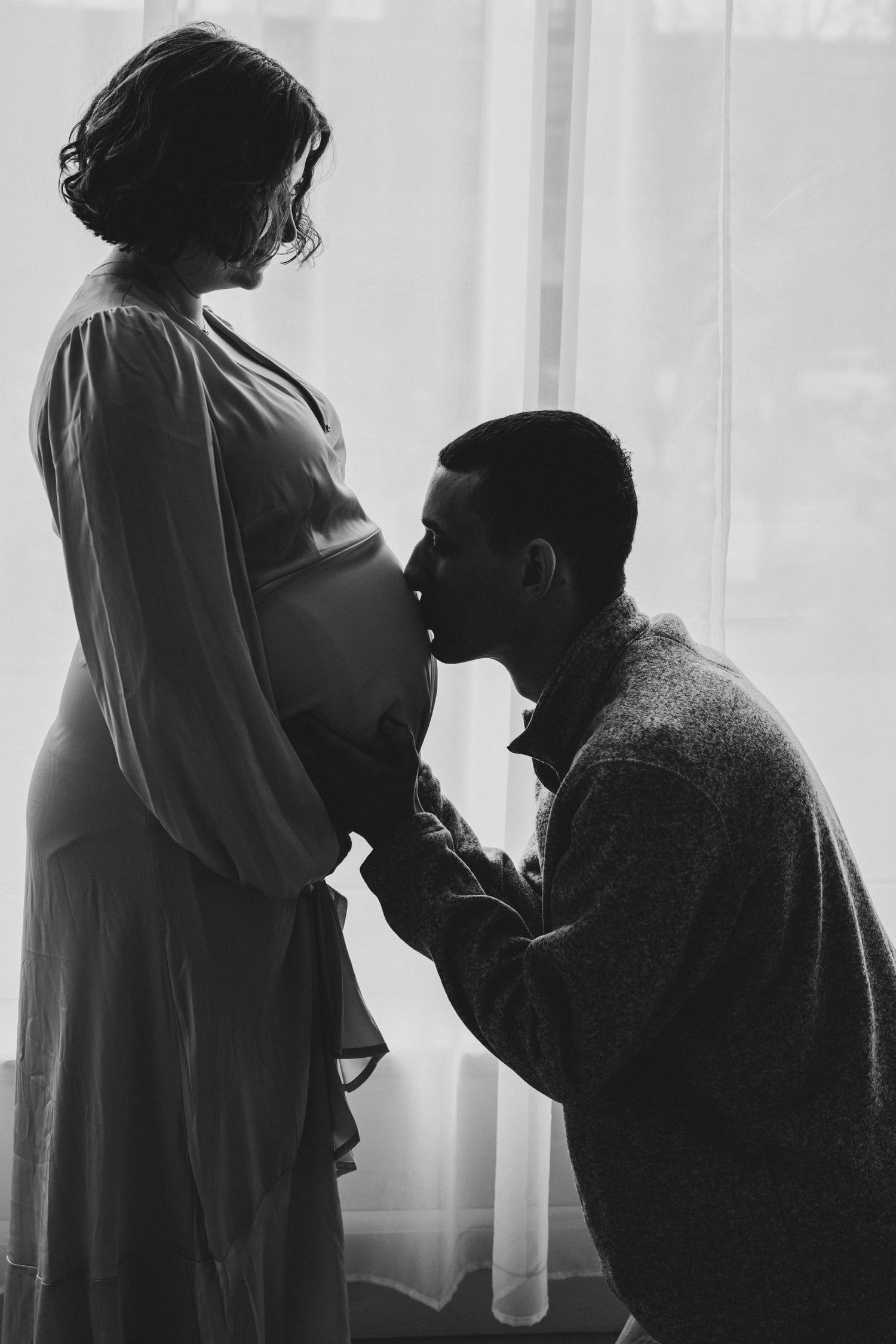 A tender silhouette of an expecting couple by a window, with the partner gently kissing the pregnant belly.