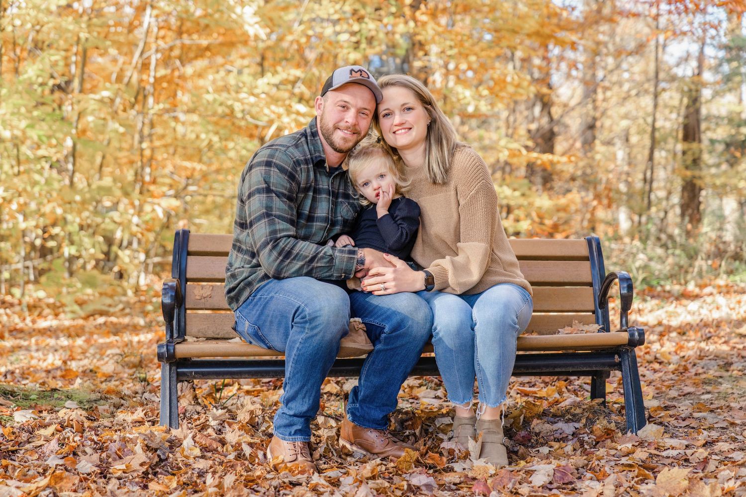 A couple sits together on a wooden bench surrounded by vibrant autumn leaves in a park setting.