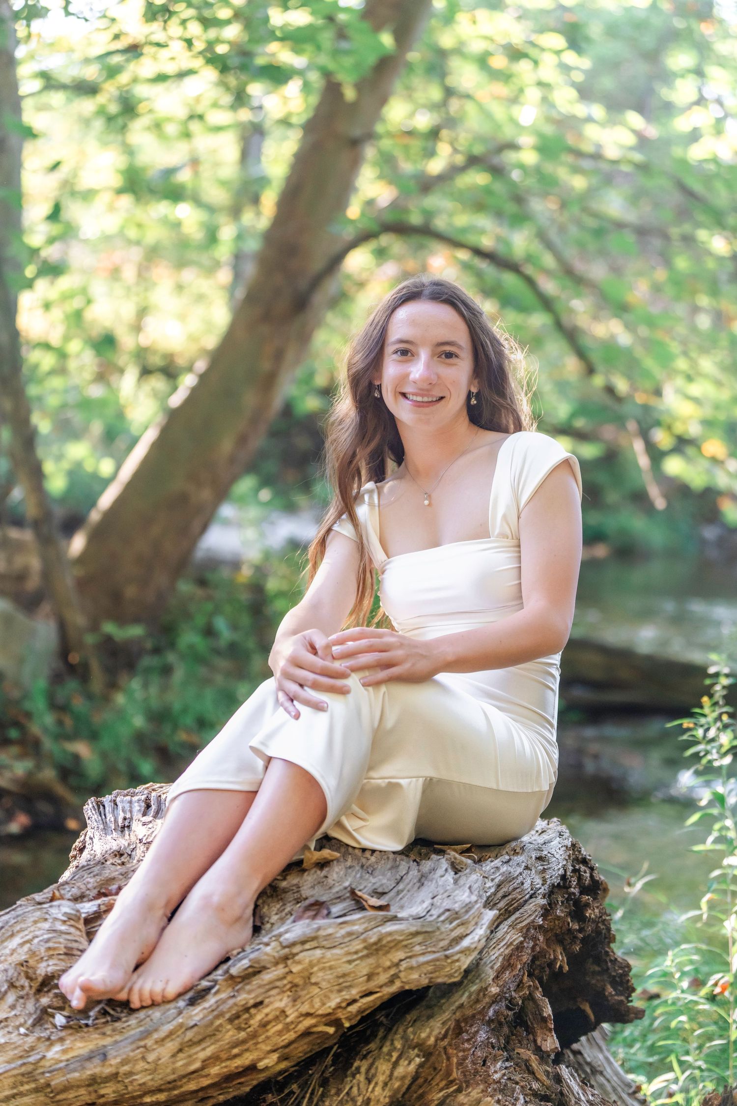 A person in a white dress sits casually on a fallen log in a sunlit forest setting.