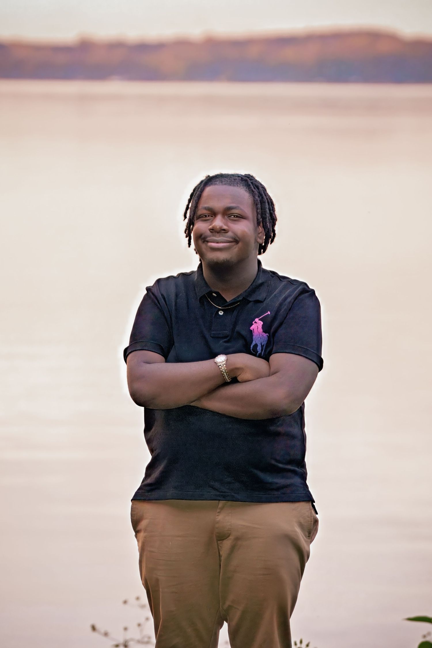 Person in black polo shirt and brown pants poses outdoors during sunset with arms crossed and friendly expression.