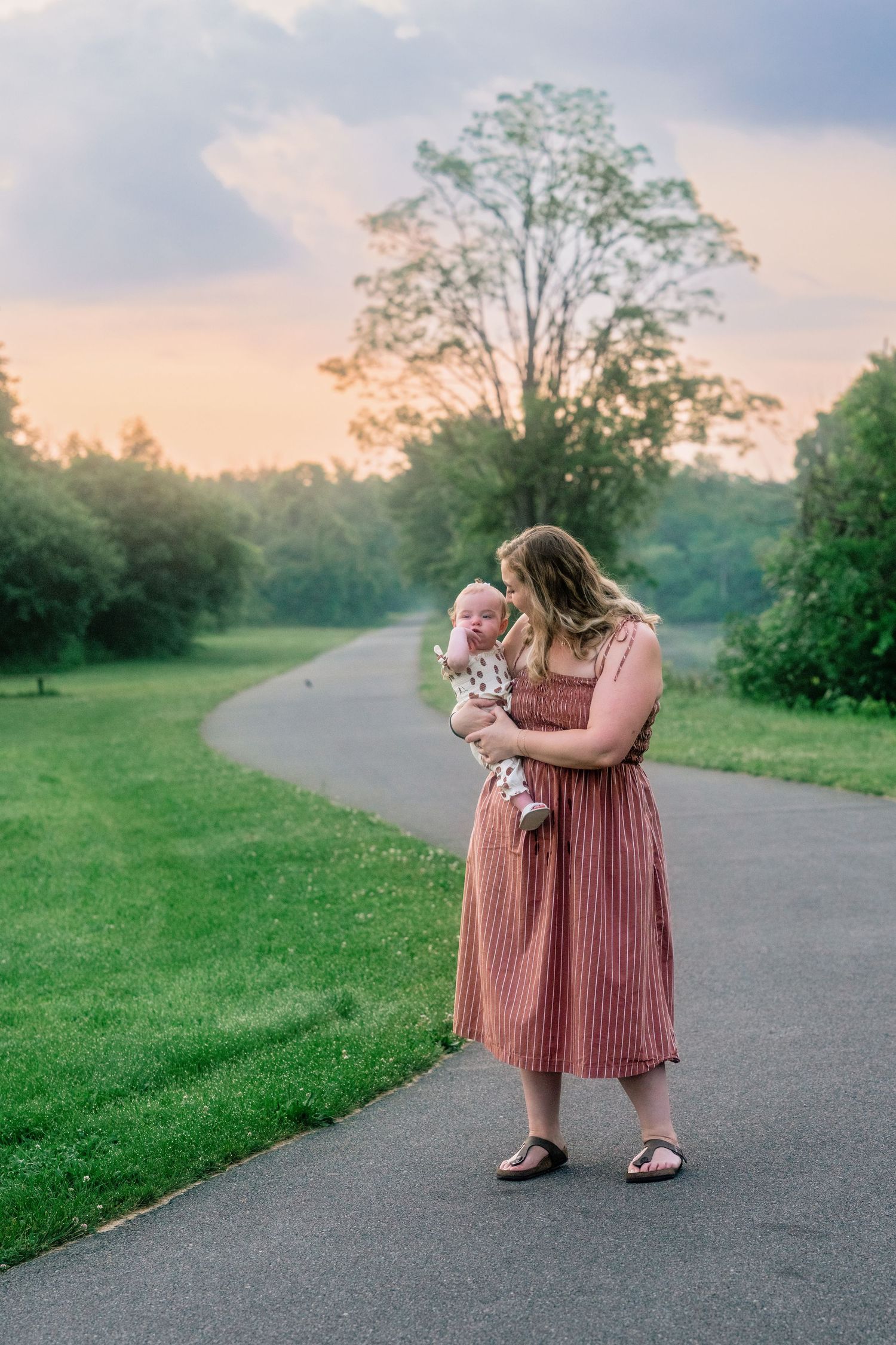 A figure in a dusty pink dress crouches down on a winding path through green grass at sunset.