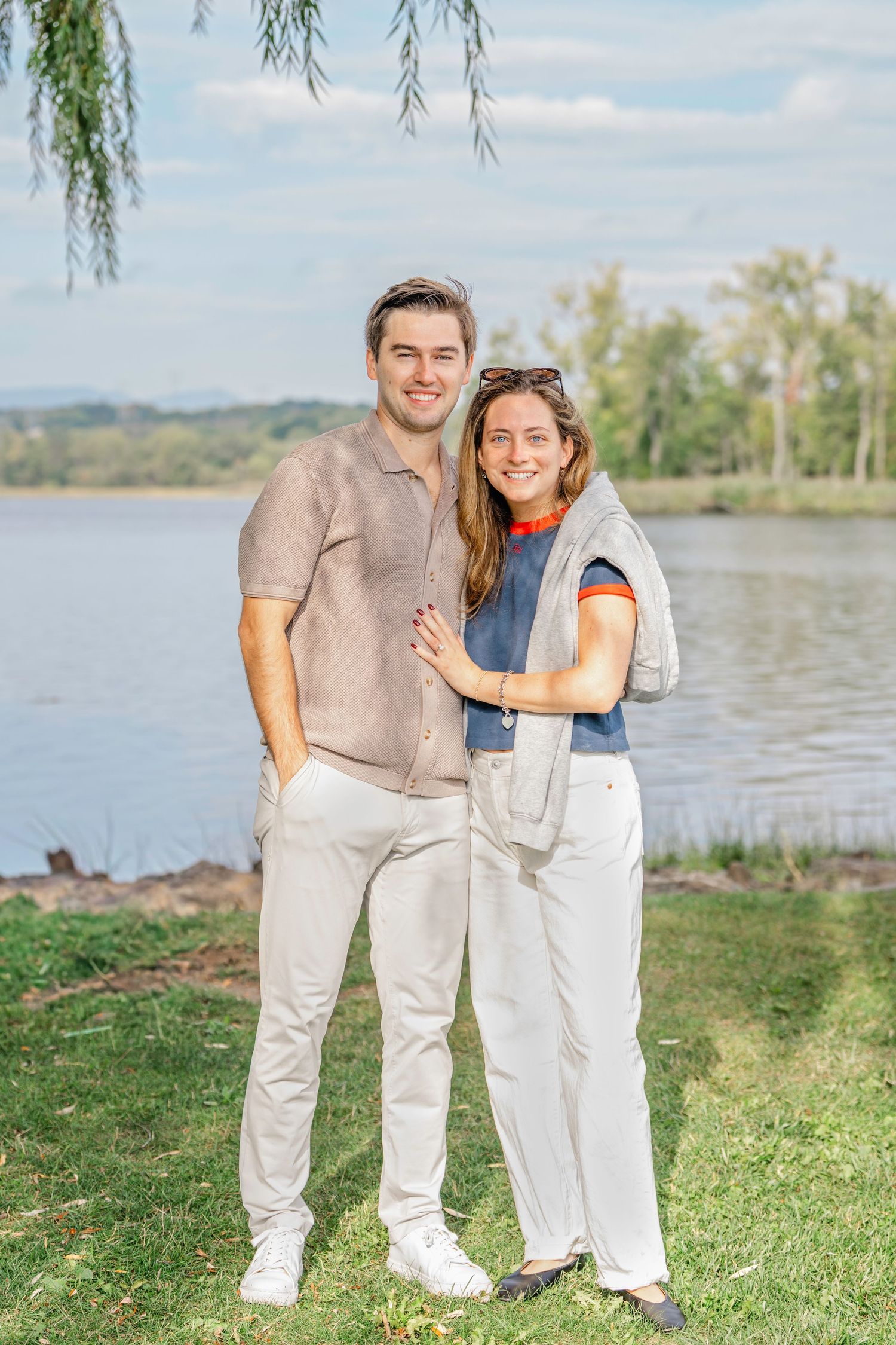 A couple in casual attire stands together on grassy lakeside with willow trees and water in the background.