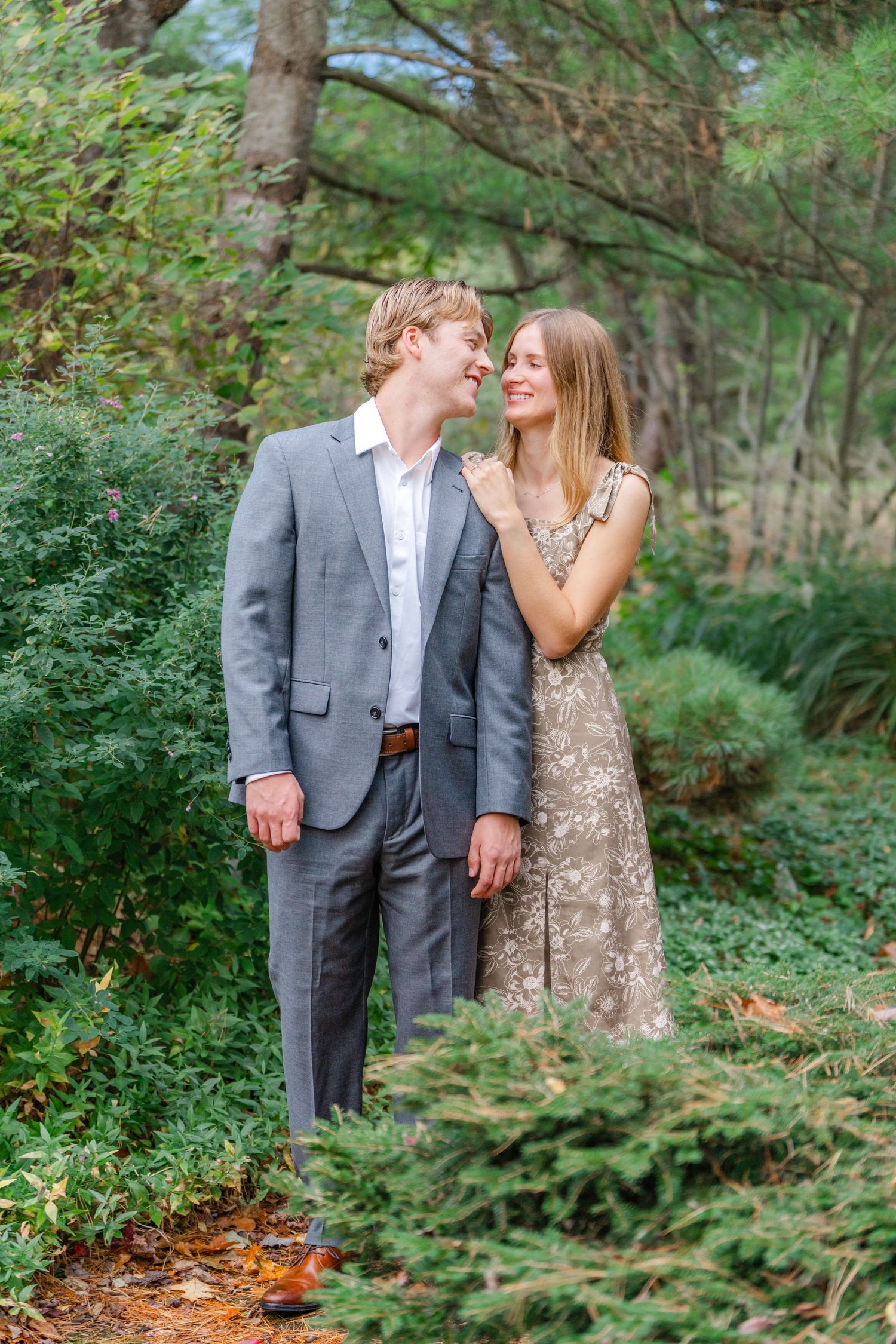A romantic couple in formal attire embraces while standing on a wooded forest path surrounded by lush greenery.