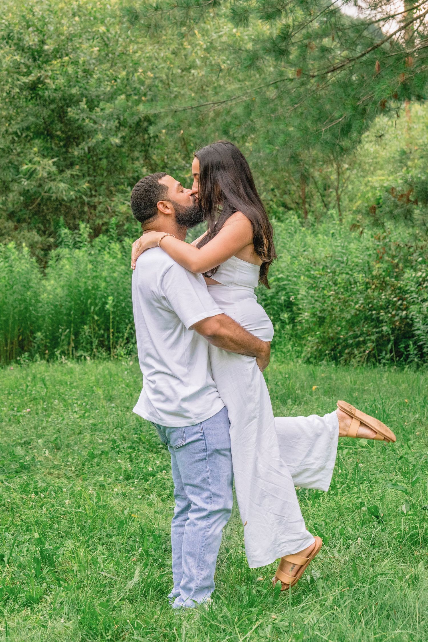 A romantic couple embraces in a joyful moment outdoors, with the woman in white being lifted up against a green backdrop.