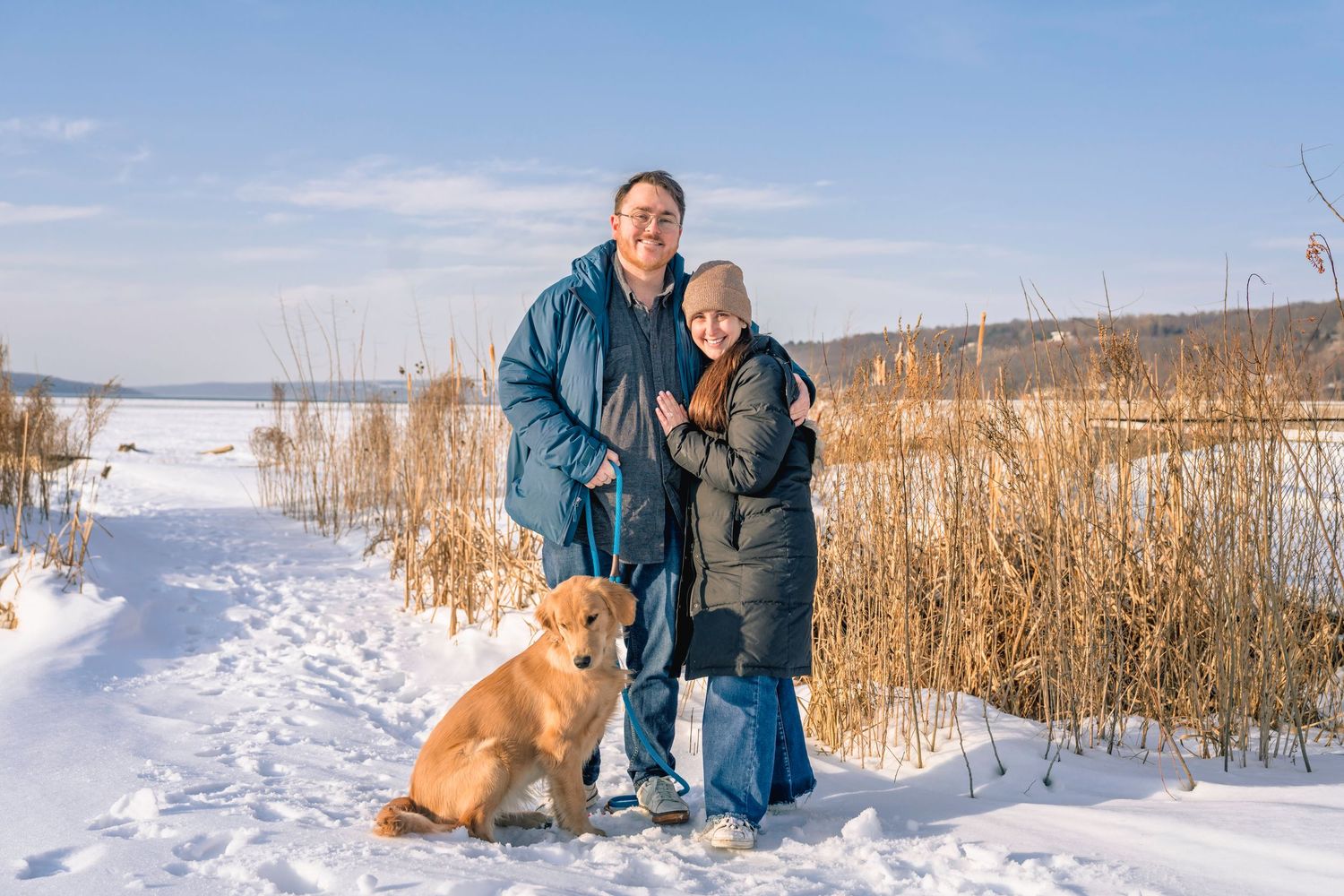 Couple posing together with golden retriever dog on snowy winter trail near frozen lake.