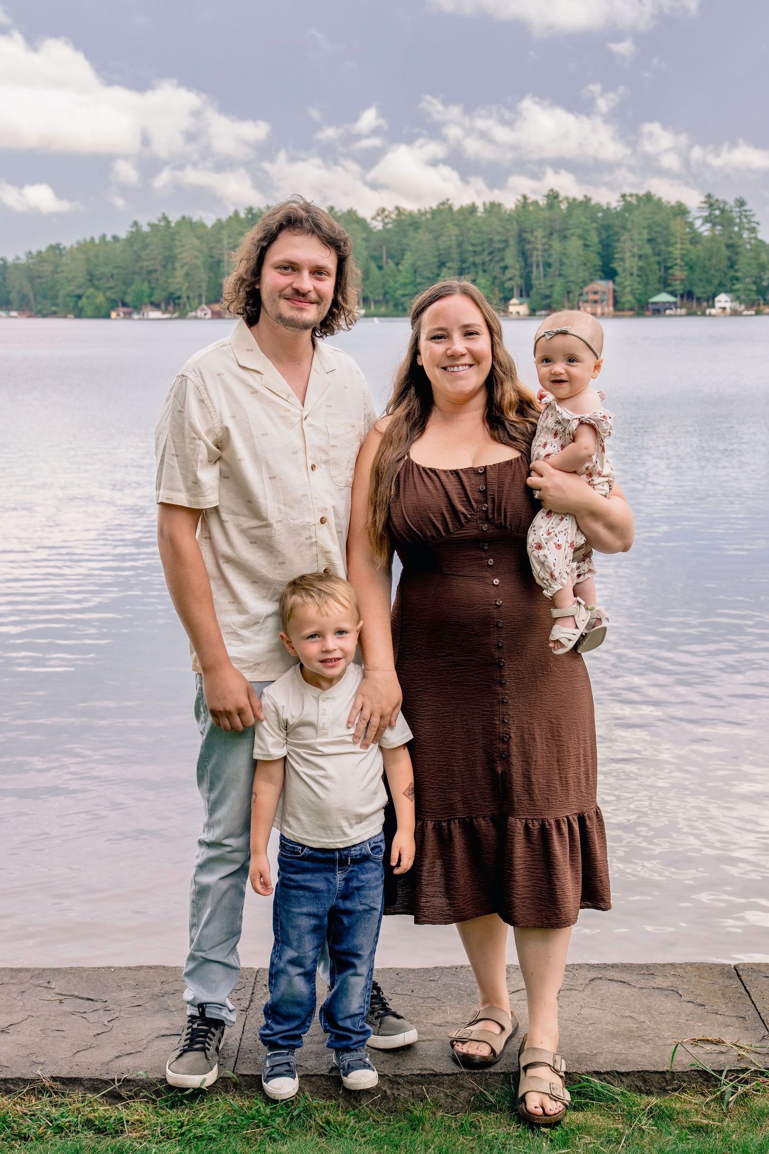 A family poses together by a serene lake with trees and houses visible across the water on a sunny day.