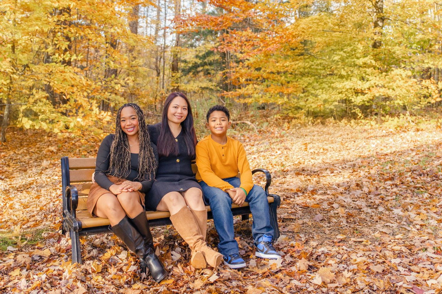 Three people sitting on a wooden bench surrounded by golden autumn leaves and fall foliage in the background.