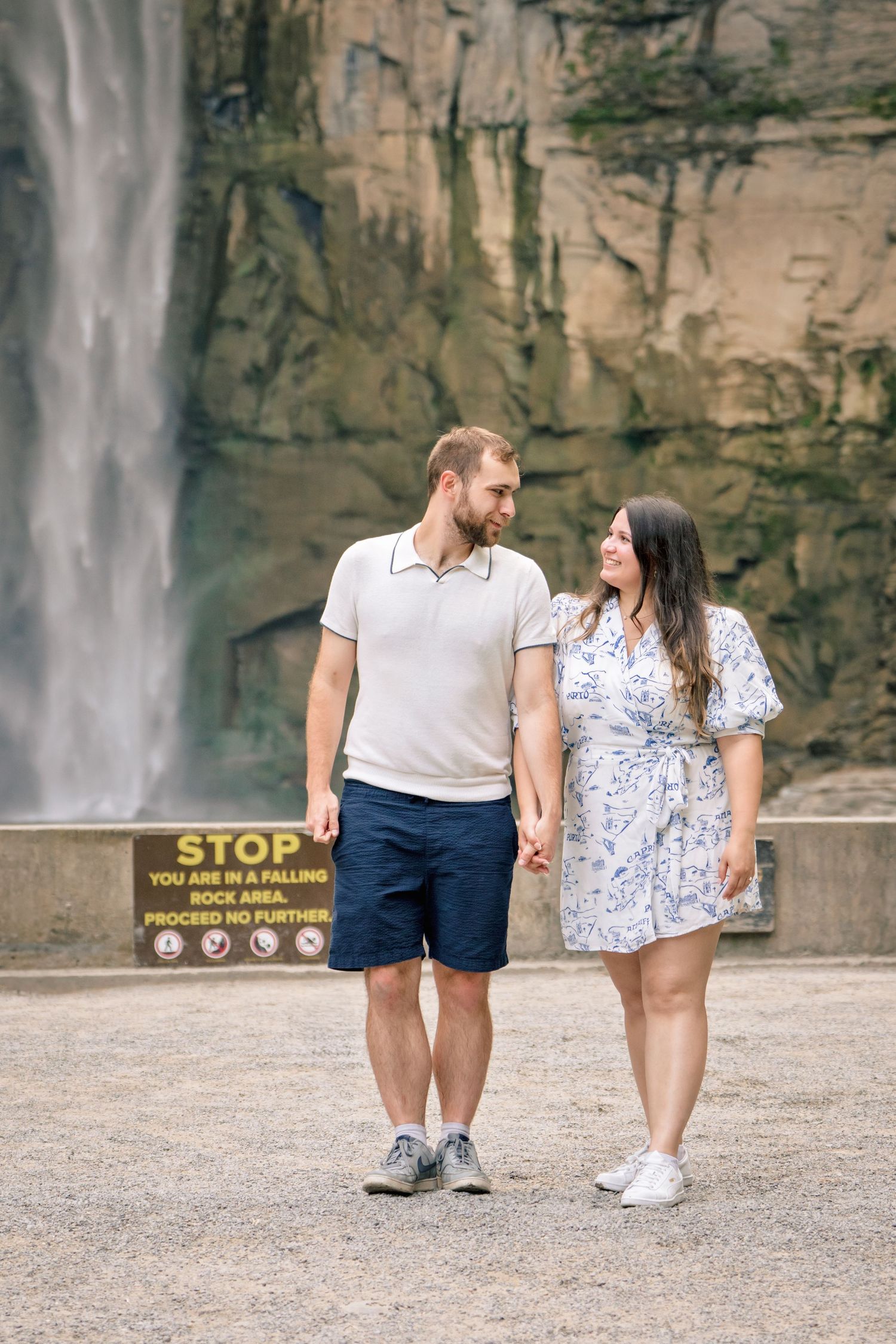 A couple takes a romantic stroll in front of a dramatic waterfall with warning sign visible in the foreground.
