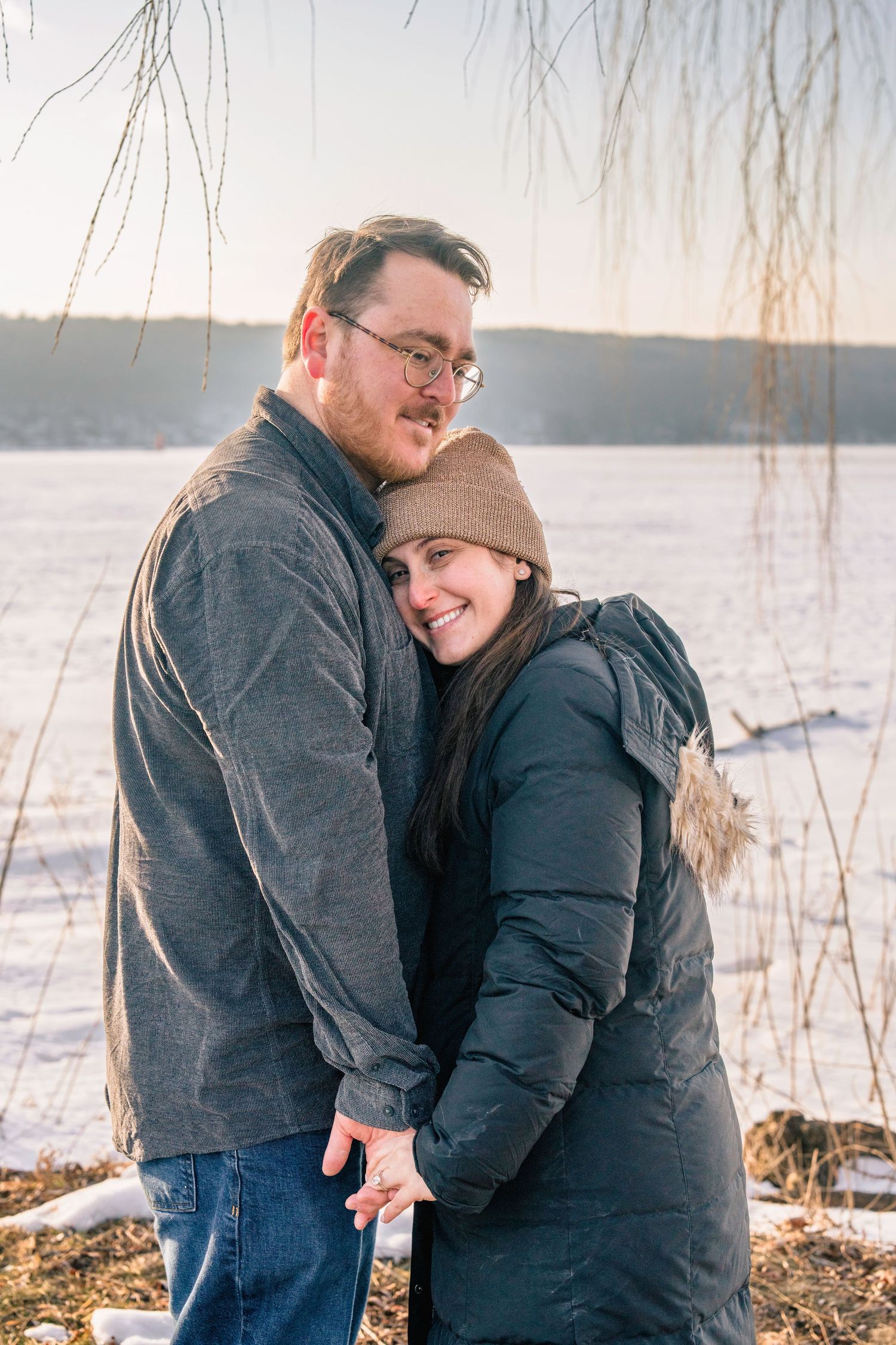 Couple embracing outdoors in winter, woman in black puffer jacket and tan beanie smiling, snowy frozen lake background.