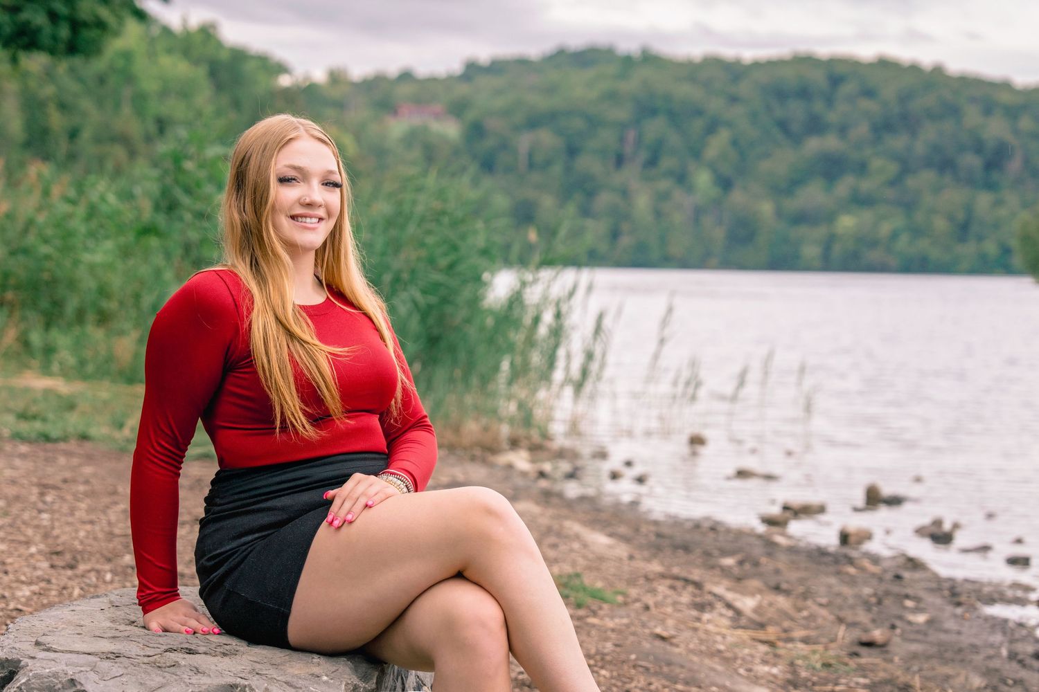 A person in a red sweater sits on a rocky shore near a lake with mountains and trees in the background. Senior photography.