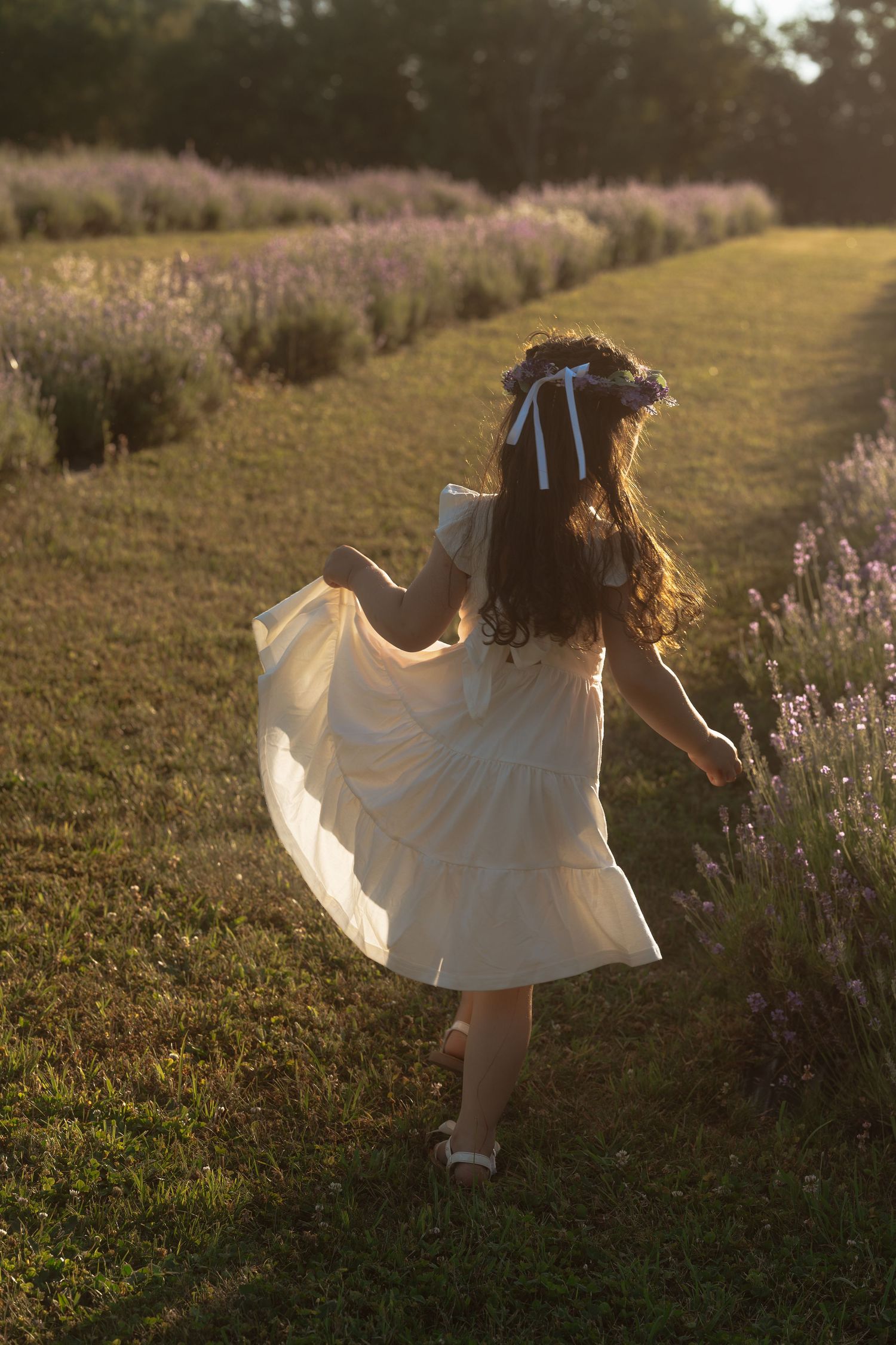 A little Girl in a flowing white dress and bow runs through a lavender field at sunset, creating a dreamy summer scene.