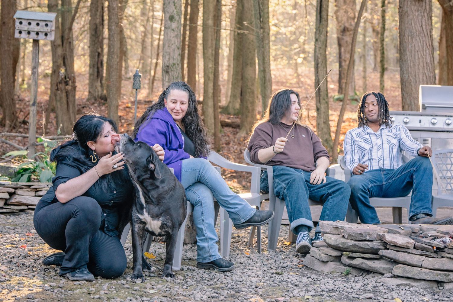 A group of friends relax and laugh together around a campfire with their dog in a wooded forest setting.