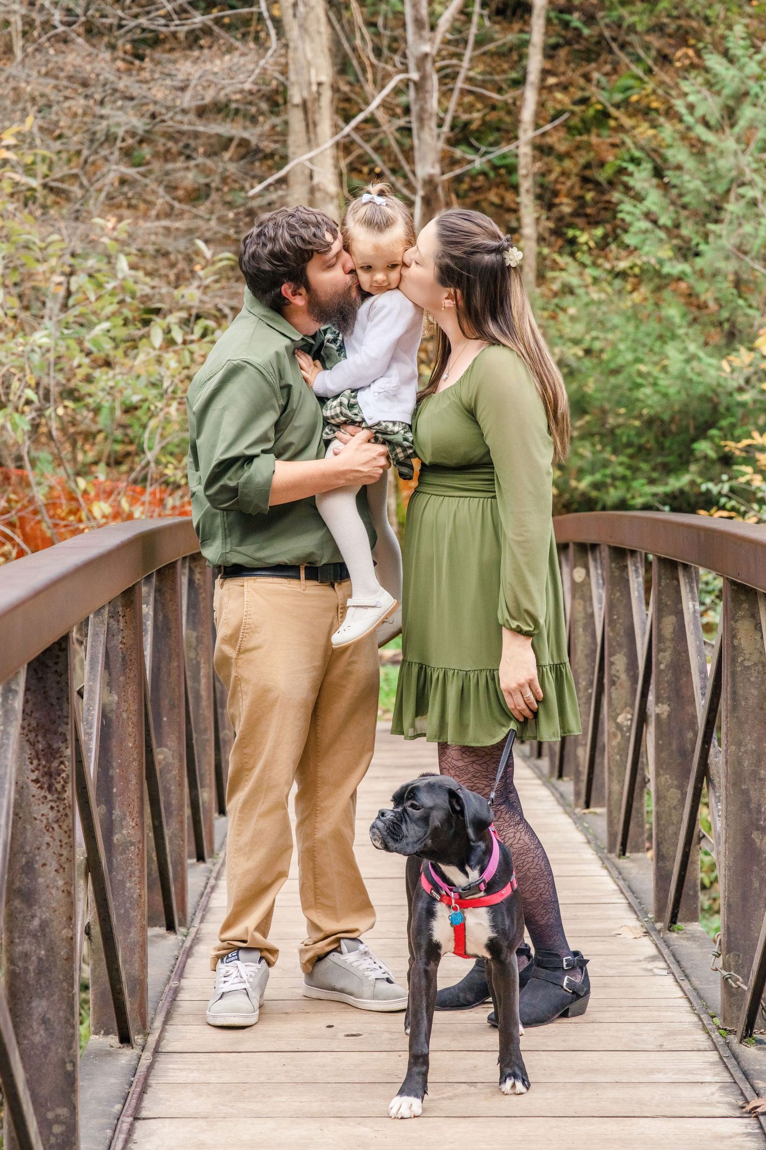 Family stands on wooden bridge with their black and white dog wearing American flag bandana in autumn forest setting.