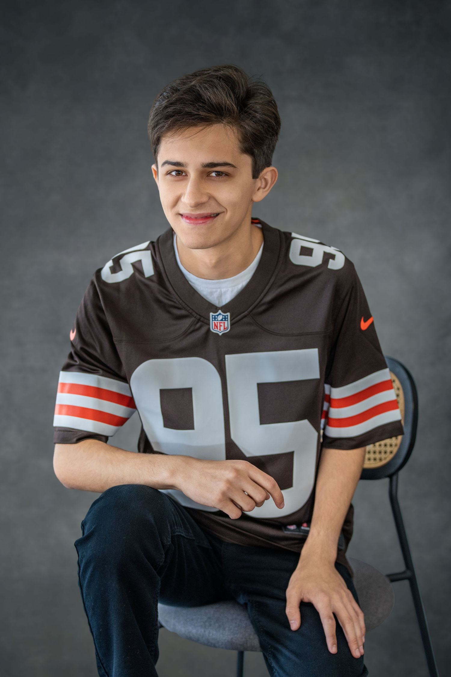 High school senior wearing a brown NFL jersey number 95 poses in front of a gray backdrop. Senior photos.