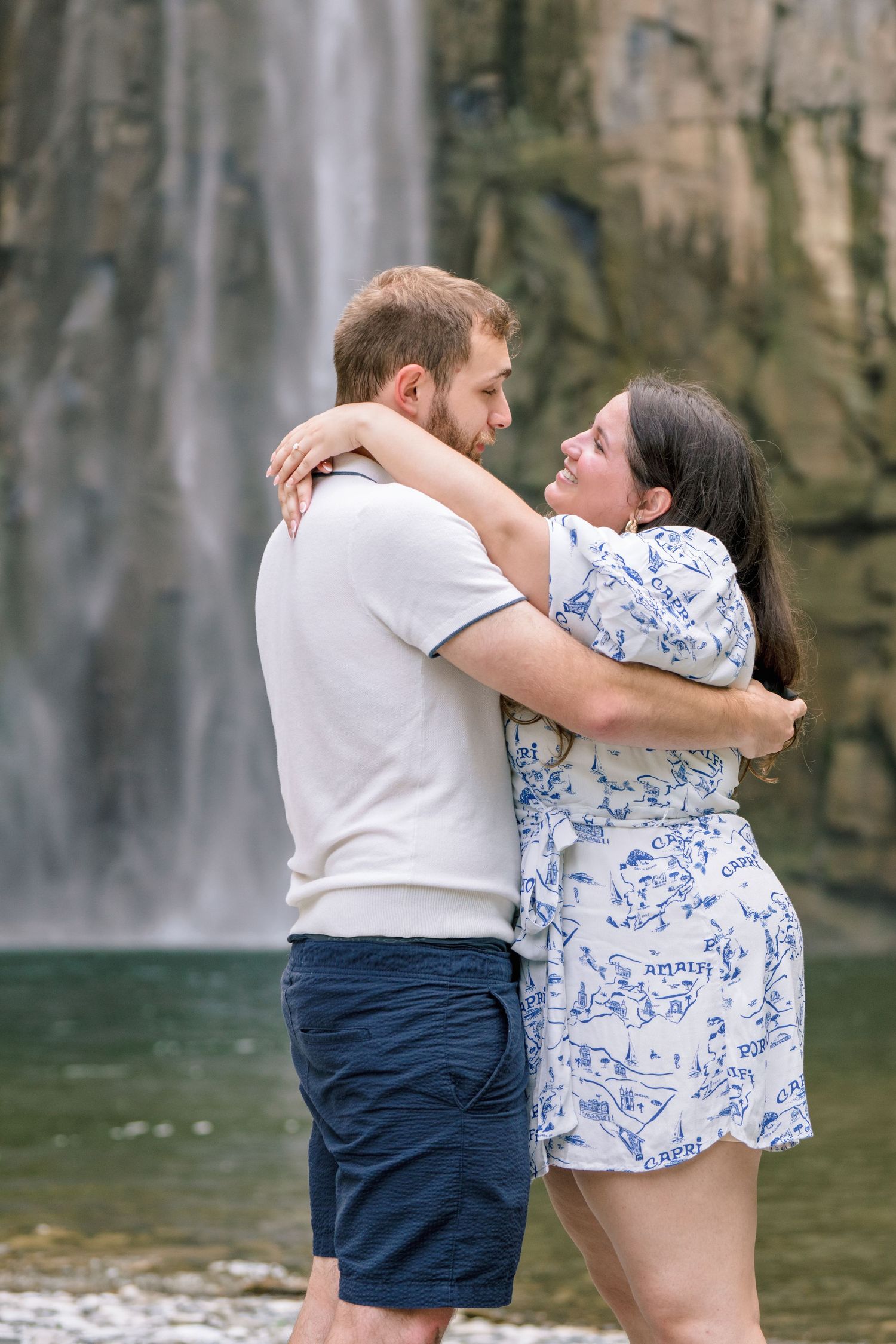 Two people share an intimate embrace while standing near a waterfall with towering rock cliffs in the background.