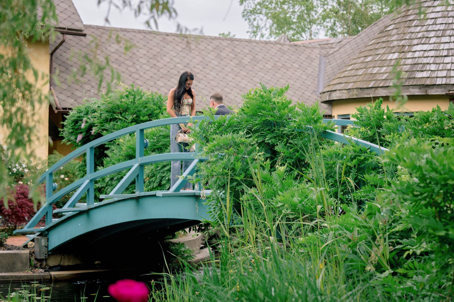 A wooden garden bridge arches over a lush pond surrounded by vibrant green plants and flowers in a tranquil setting.