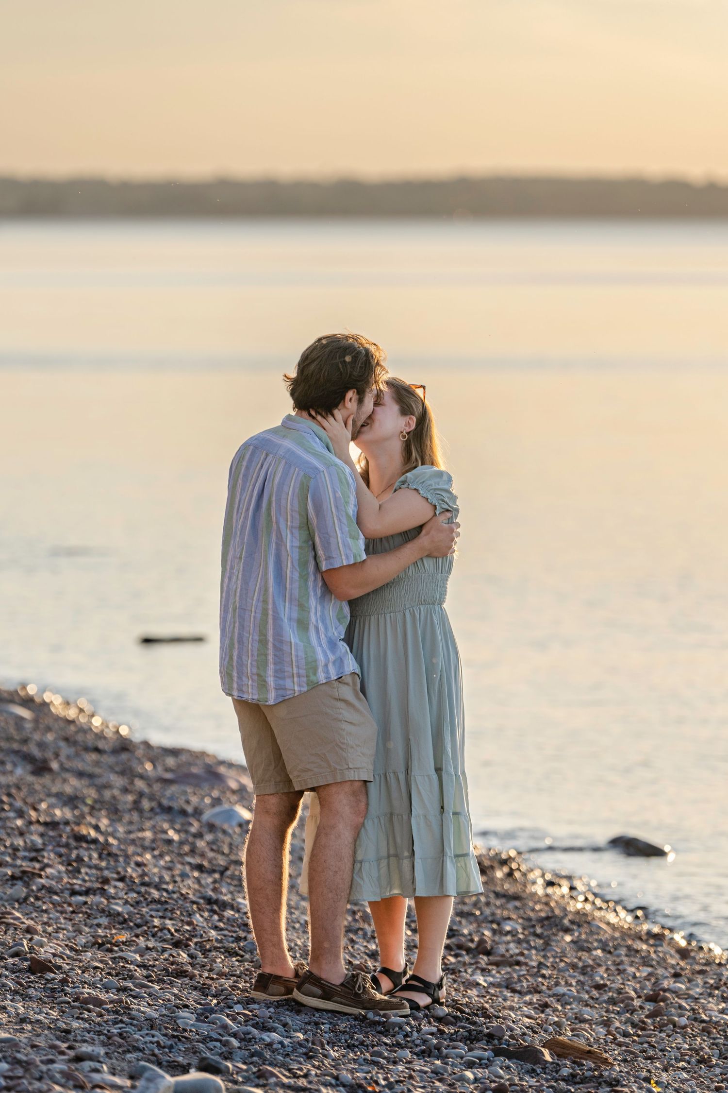 A couple shares a tender moment at sunset on a rocky beach shore with calm waters in the background.