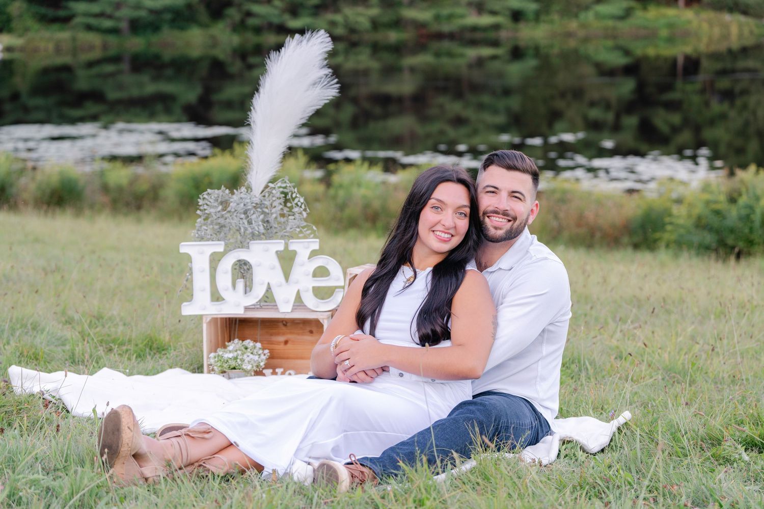 A couple poses for engagement photos on grass with decorative 'LOVE' letters nearby.
