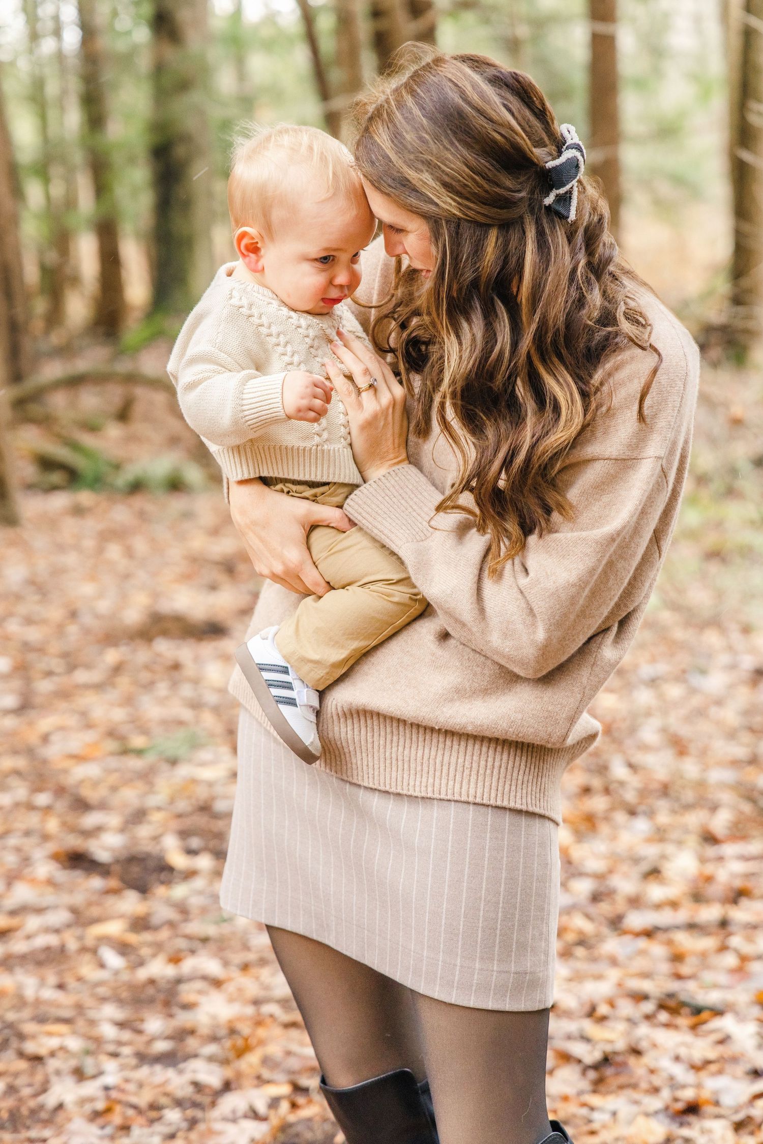 Parent and baby wearing matching beige sweaters embrace while standing among fallen autumn leaves in a forest.