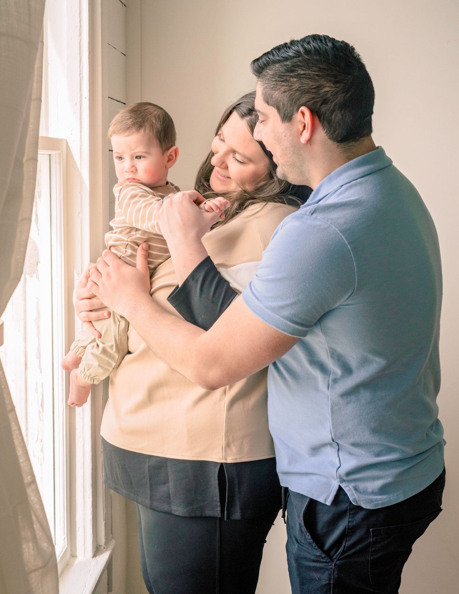 A happy family of three stands by a window, parents lovingly holding their infant baby.