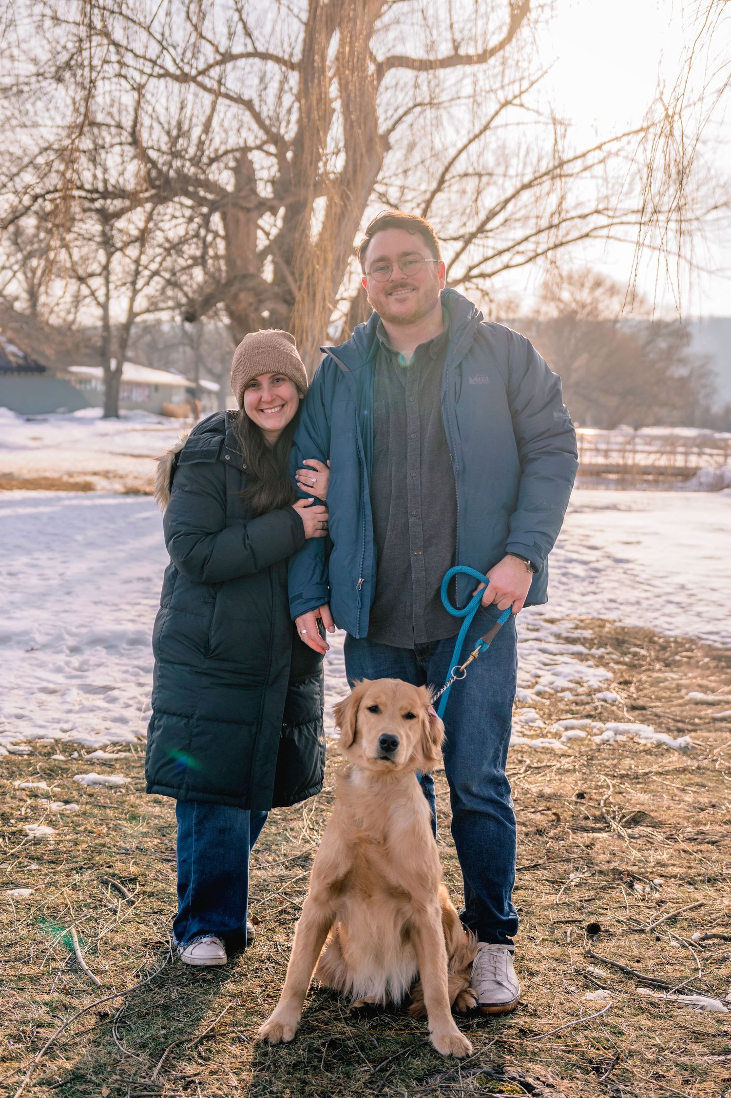 Smiling couple standing with golden retriever on leash in snowy park during golden hour.