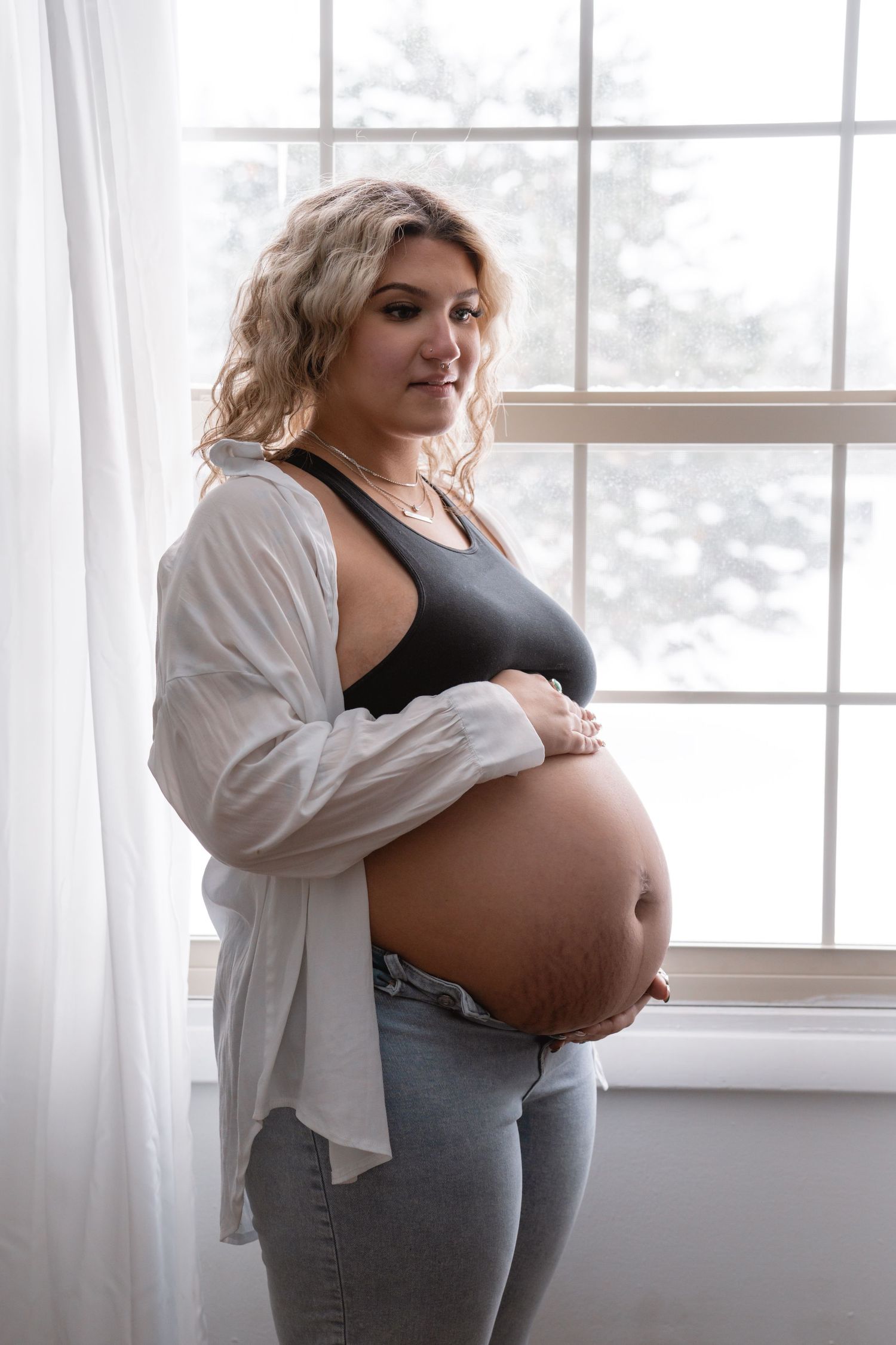 Expectant mother stands by window in casual clothing while cradling her pregnant belly in a tender maternity pose.
