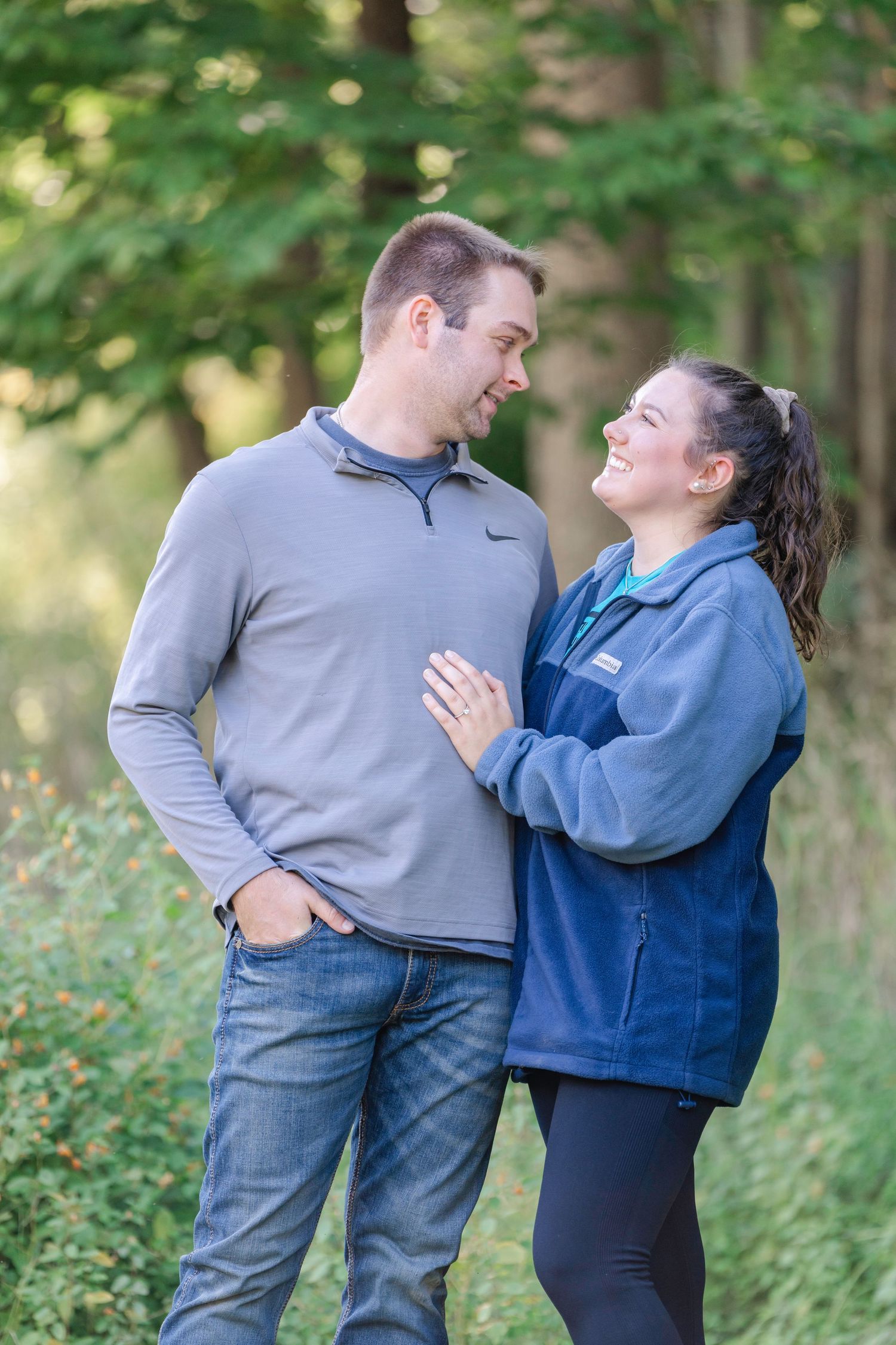 A couple shares a joyful moment together outdoors in a wooded setting wearing casual blue and grey clothing.