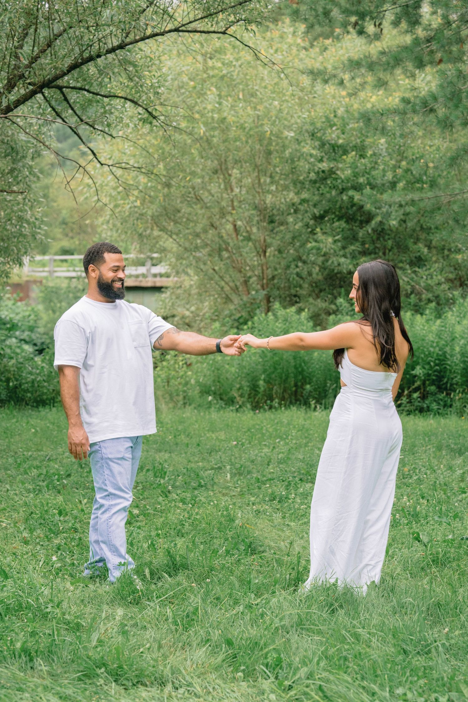 A couple in white outfits playfully interacting in a lush green outdoor setting during a photo session.