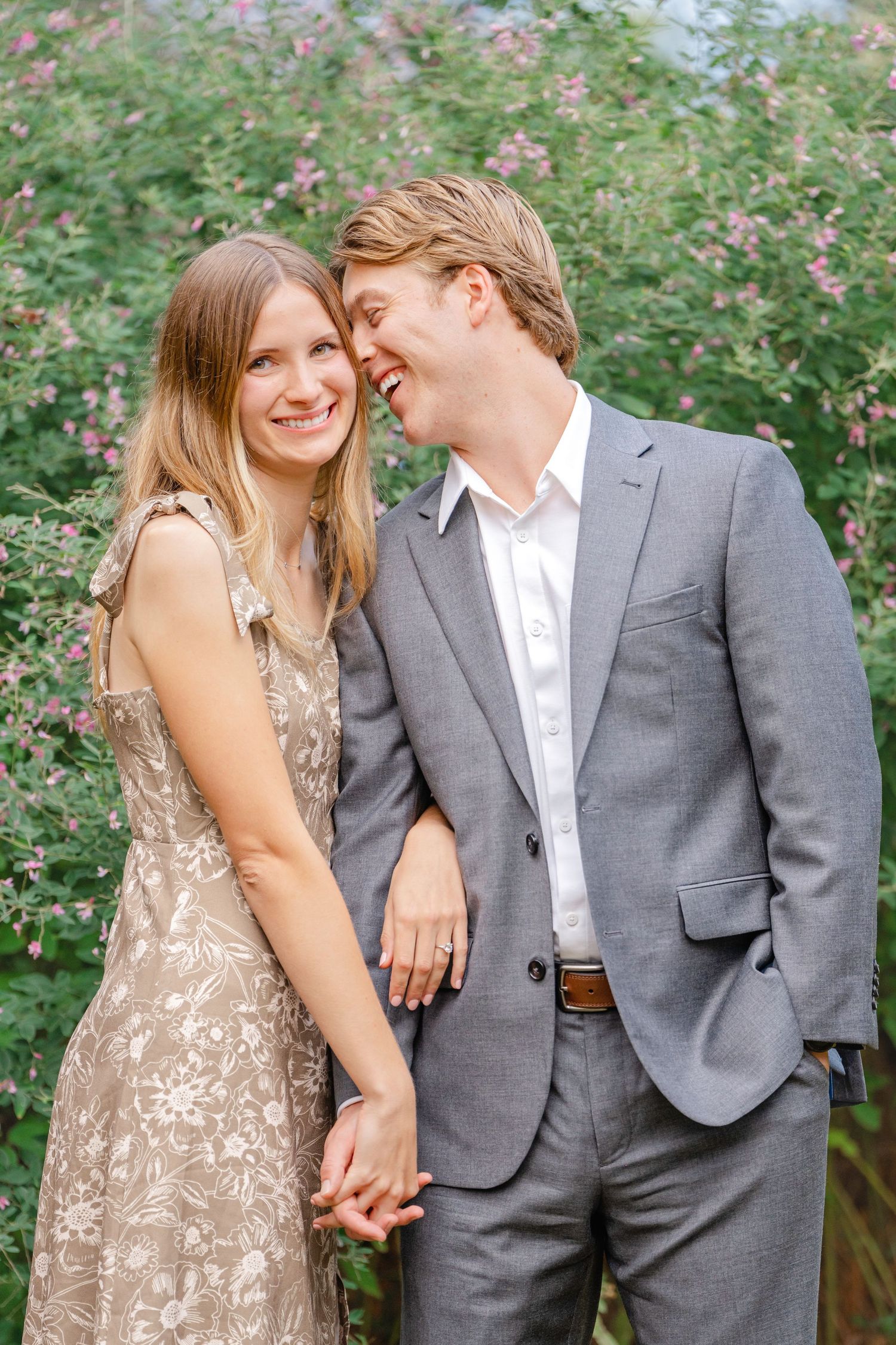 A couple shares a romantic moment outdoors wearing formal attire, with flowering bushes in the background.