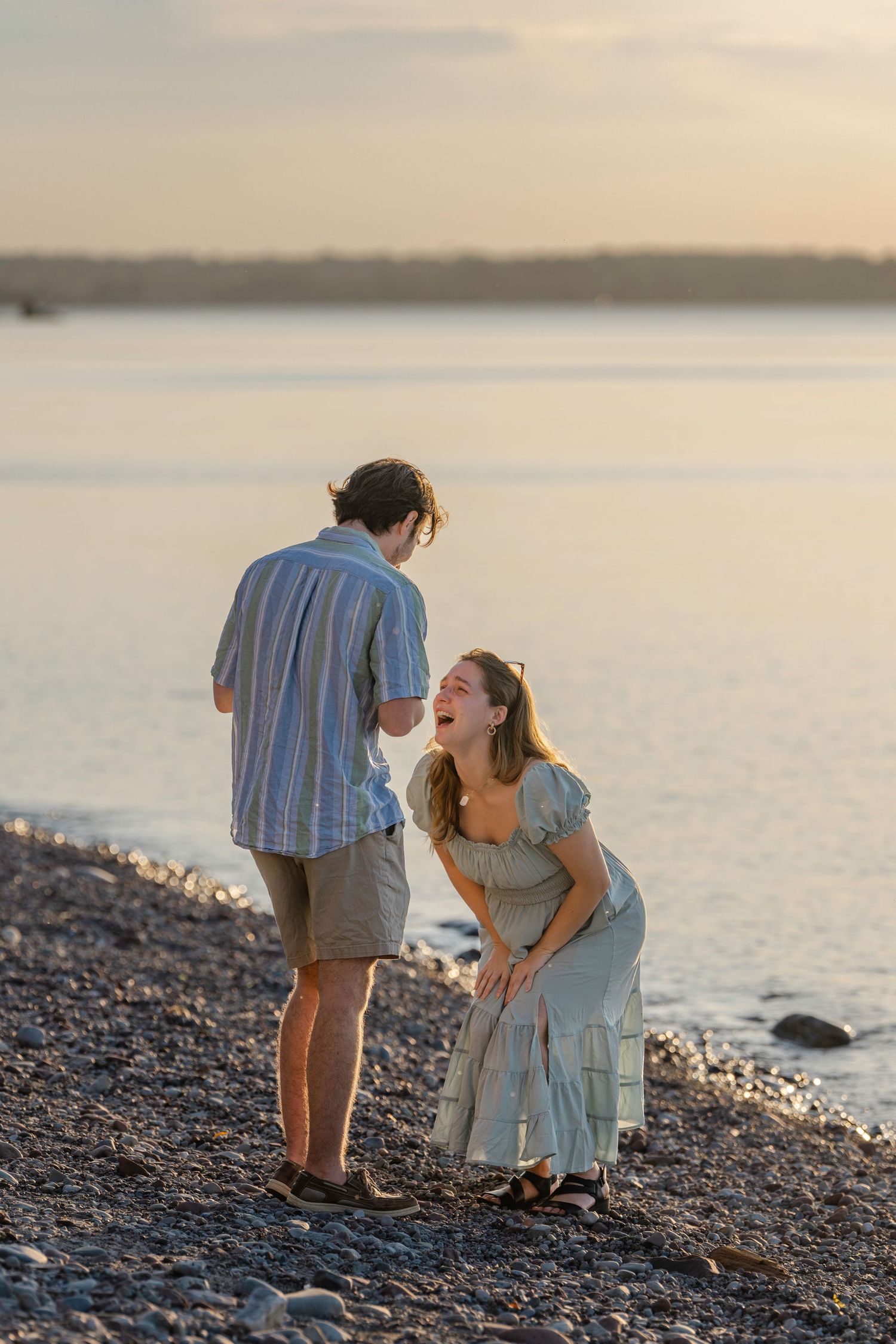 Two people embrace while sitting on a pebbled beach at golden hour as the sun sets over tranquil waters.