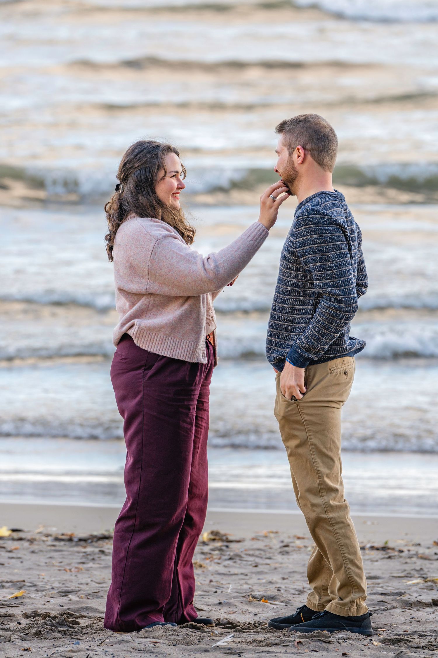 A couple shares a playful moment together on a sandy beach during what appears to be an engagement photoshoot.