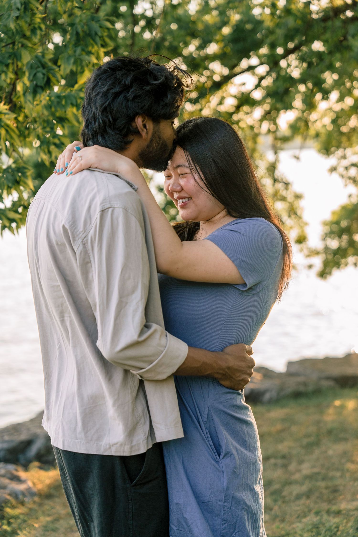 A romantic couple embraces outdoors under dappled sunlight and green tree branches in a tender moment.