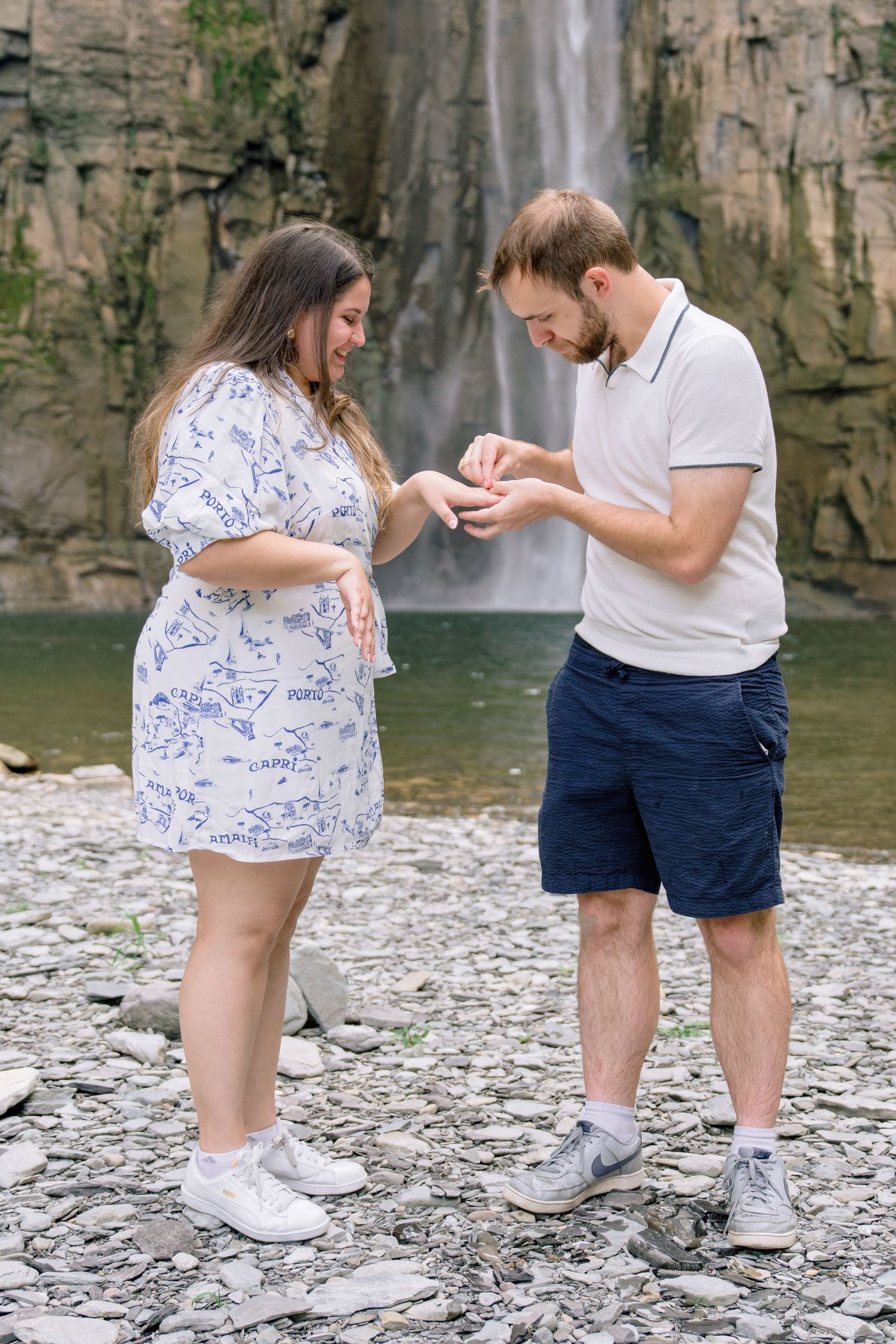 A couple kneels on rocky ground near a waterfall during what appears to be a proposal moment.