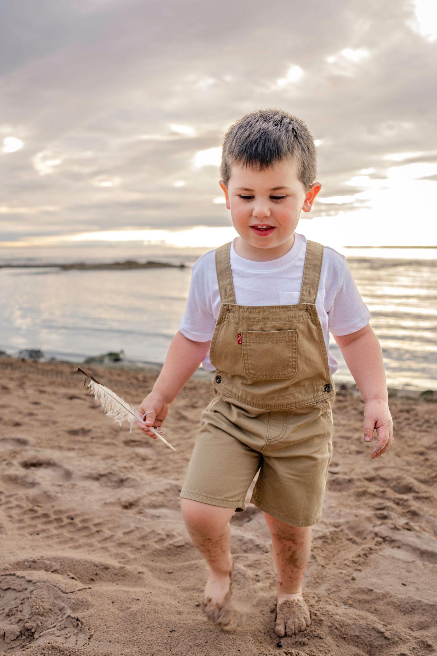 A toddler in brown overalls plays on a sandy beach at sunset with waves and cloudy skies in the background.