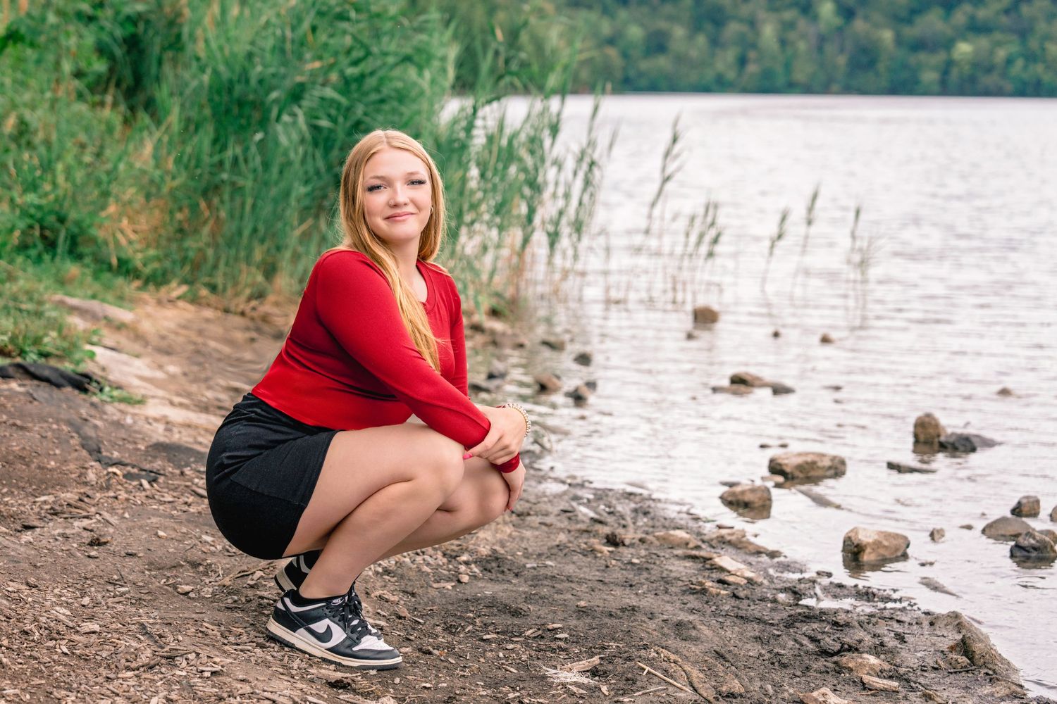 A person in a red sweater sits by a lake watching ducks swim in the water on a cloudy day. Senior photos