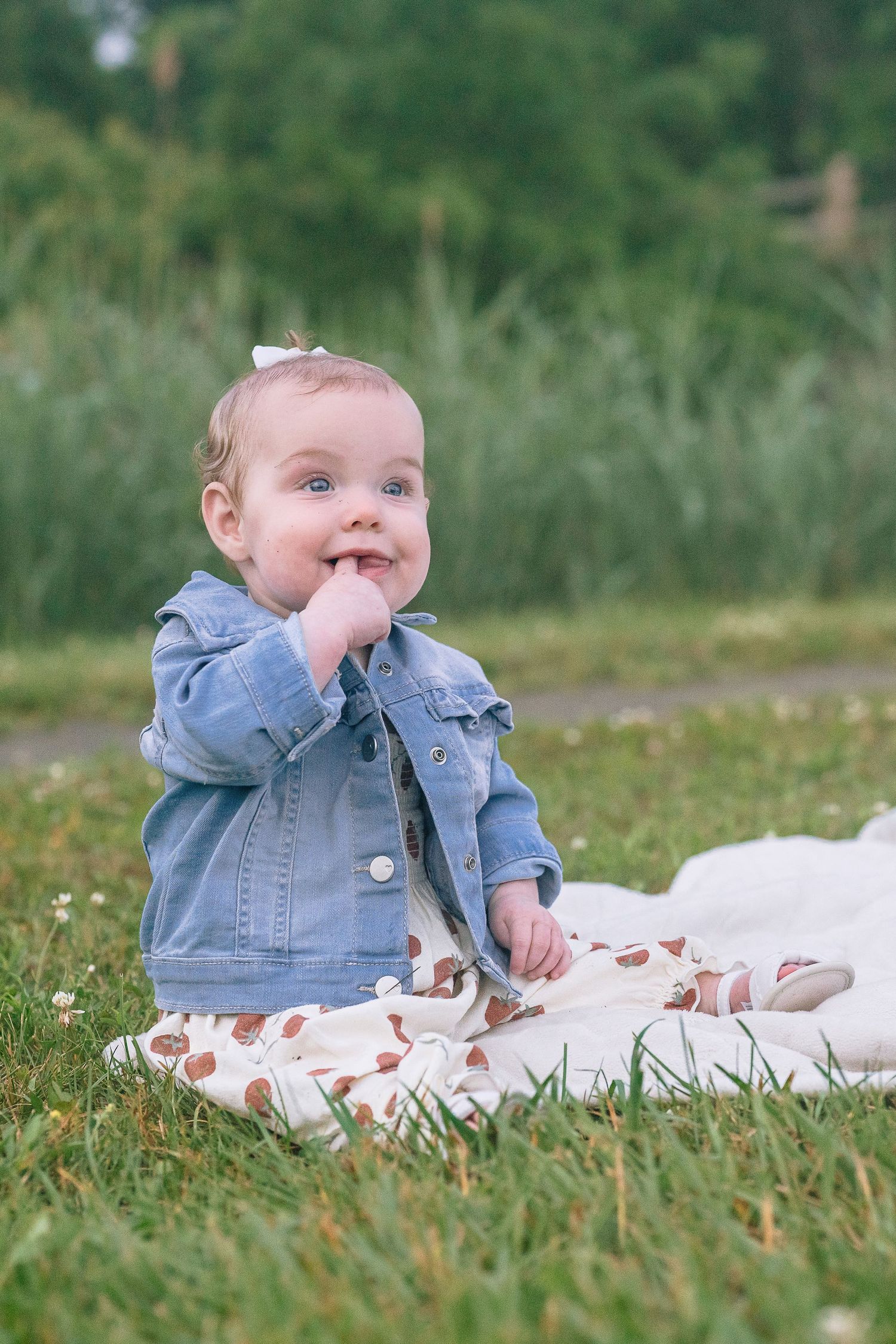 Baby in denim jacket sits on blanket in grass with a joyful expression.