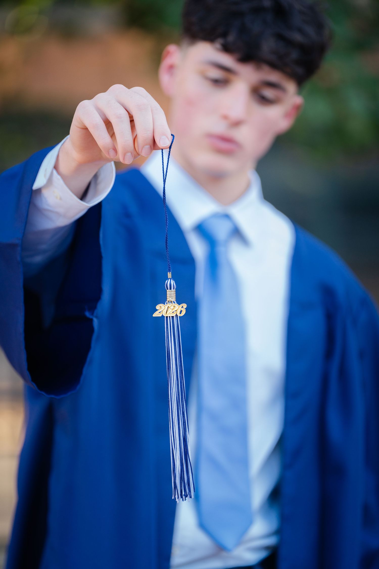 A person in a blue suit and tie holds a graduation tassel against a blurred outdoor background.