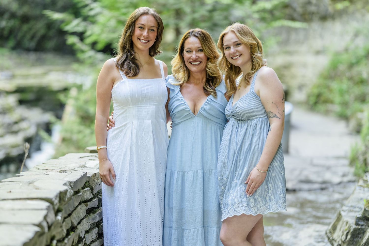 Three people in flowing pastel dresses pose together on a stone path surrounded by greenery.
