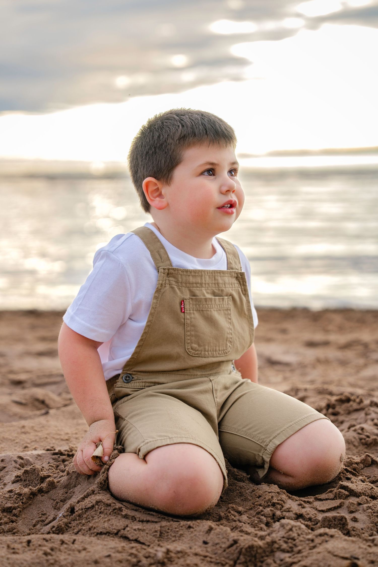 Small child wearing tan overalls and white shirt sits on sandy beach during cloudy sunset.