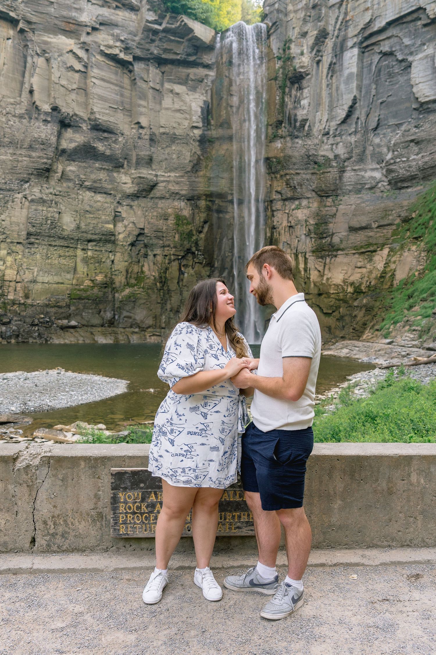 A couple sharing an emotional moment on a stone wall with dramatic cliffs and waterfall scenery behind them.