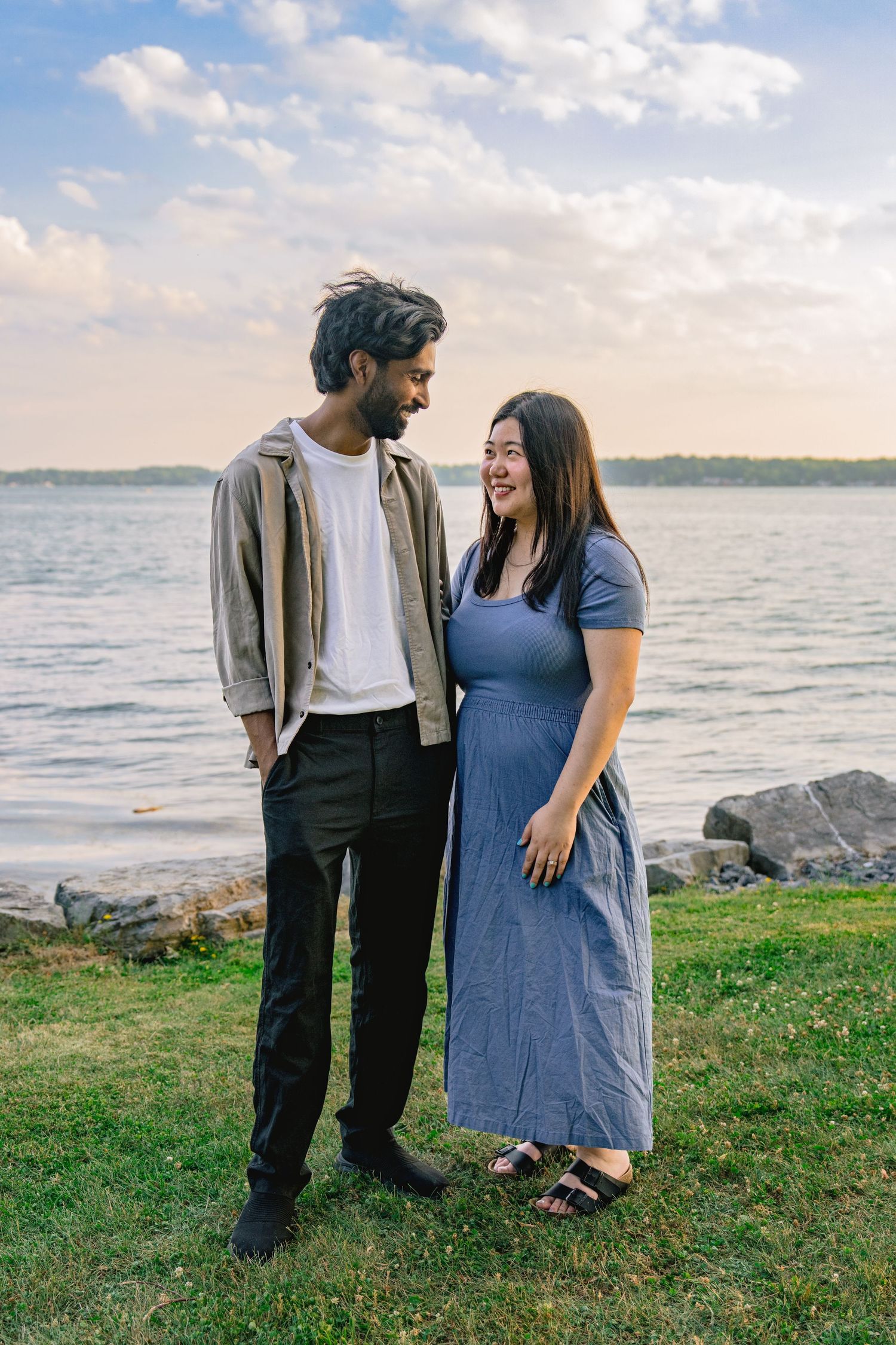 A couple stands together on a grassy shore overlooking a serene lake at sunset.