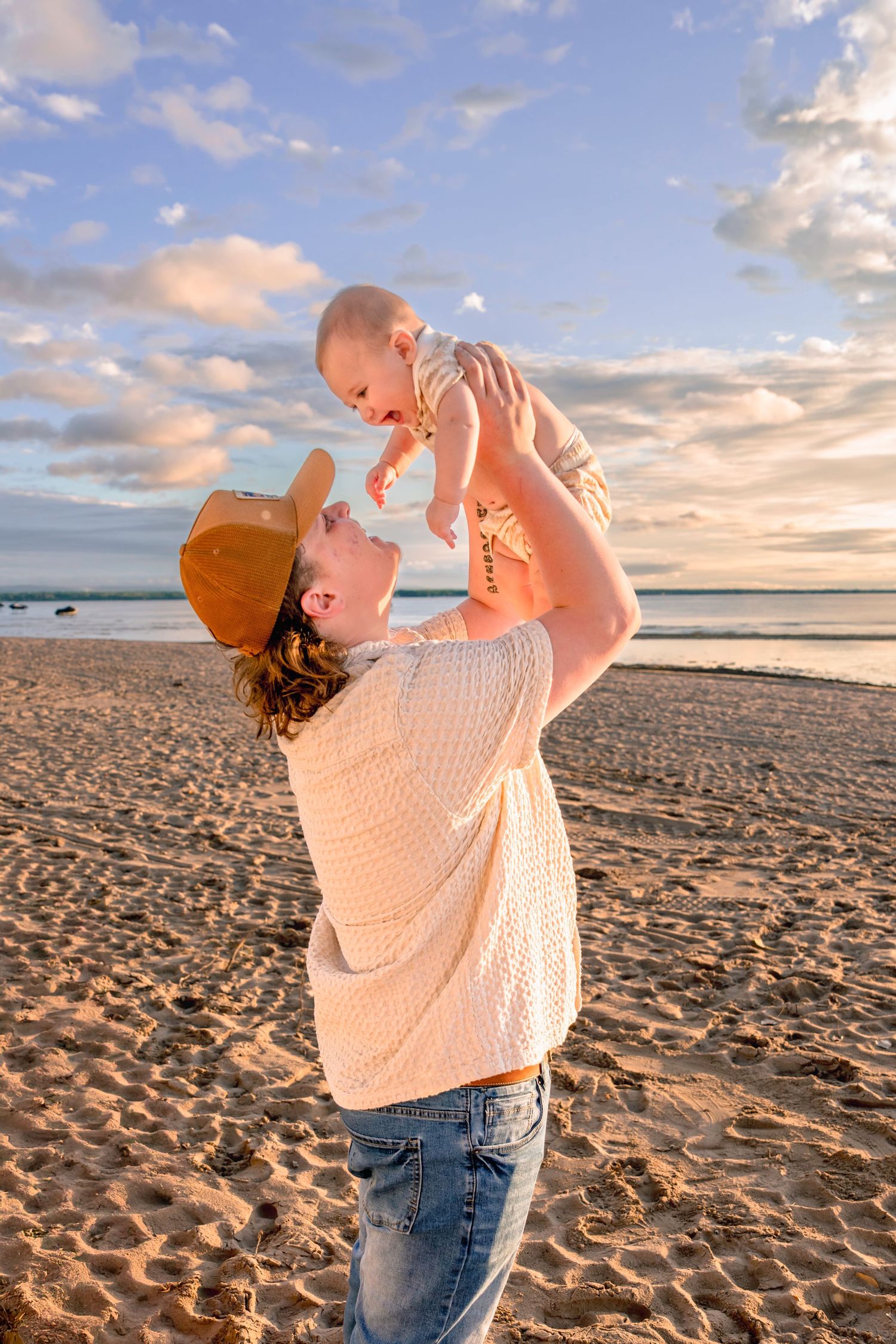 Person in white sweater lifts baby joyfully against sunset sky on beach.